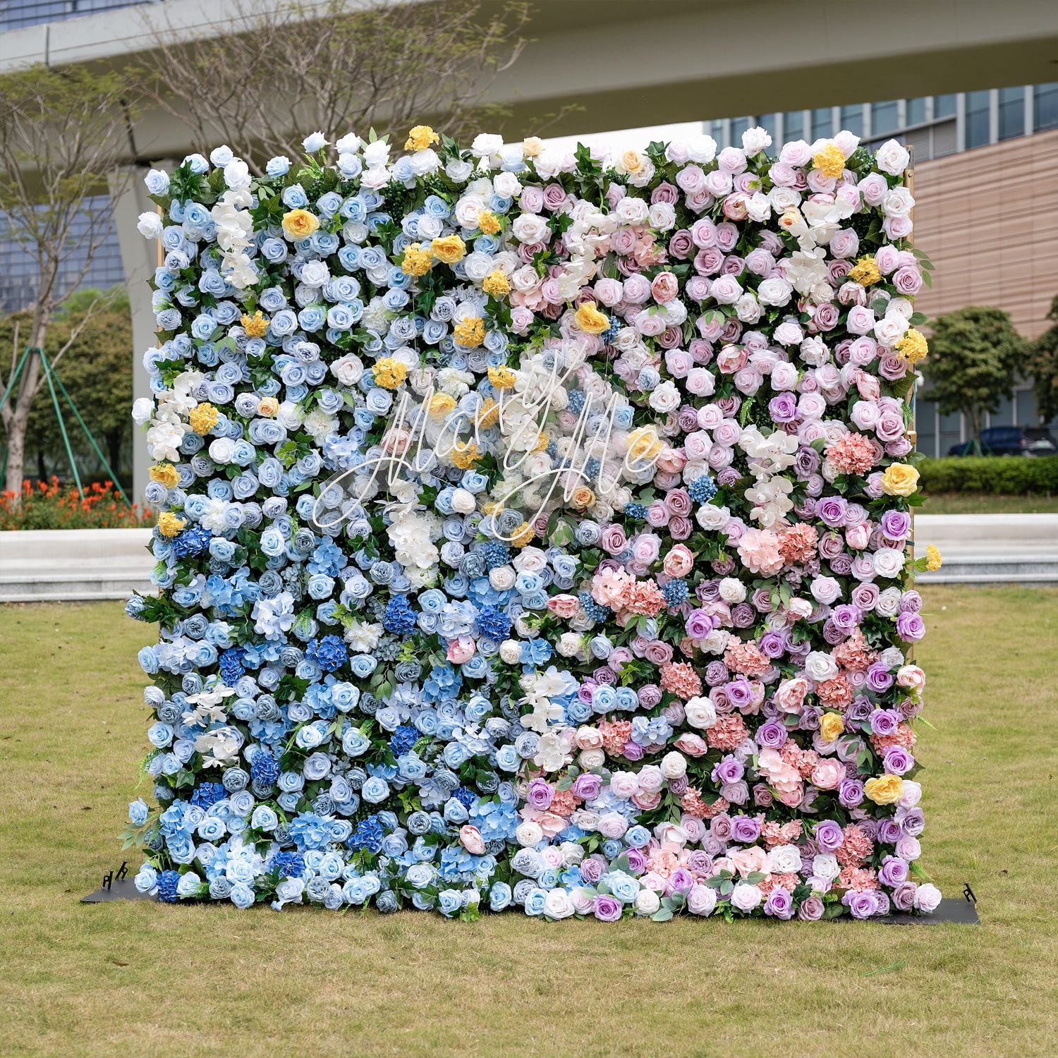 This is a stunning, ultra-dense artificial flower wall from ValarFlowers, bursting with lifelike blooms in a dreamy pastel gradient. It features sky-blue roses, creamy white roses, sunny yellow carnations, blush pink peonies, lavender roses, and lush green foliage, with the brand’s elegant gold script logo at the center. This premium, reusable floral arrangement creates a romantic, showstopping focal point ideal for weddings, photo booths, bridal showers, and luxury event decor.
