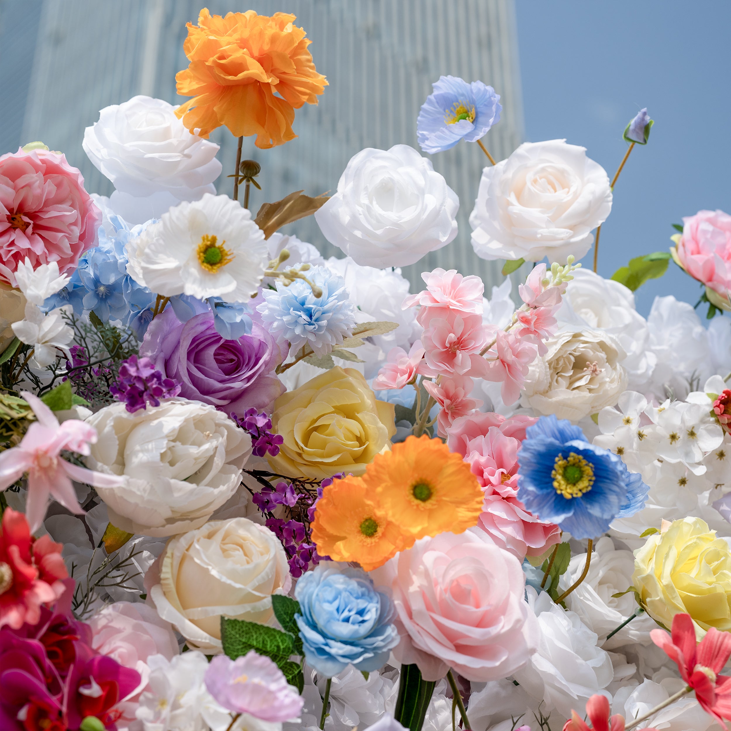 This is a lush, vibrant close-up of a premium artificial mixed-flower arrangement, brimming with lifelike blooms in a dreamy, colorful palette. It features creamy white roses, soft pink peonies, sunny yellow roses, vivid orange carnations, sky-blue poppies, lavender roses, and delicate filler flowers, set against a bright blue sky and modern glass building backdrop. The layered, realistic petals create a romantic, whimsical aesthetic, perfect for wedding backdrops, event decor, and photo booth styling.
