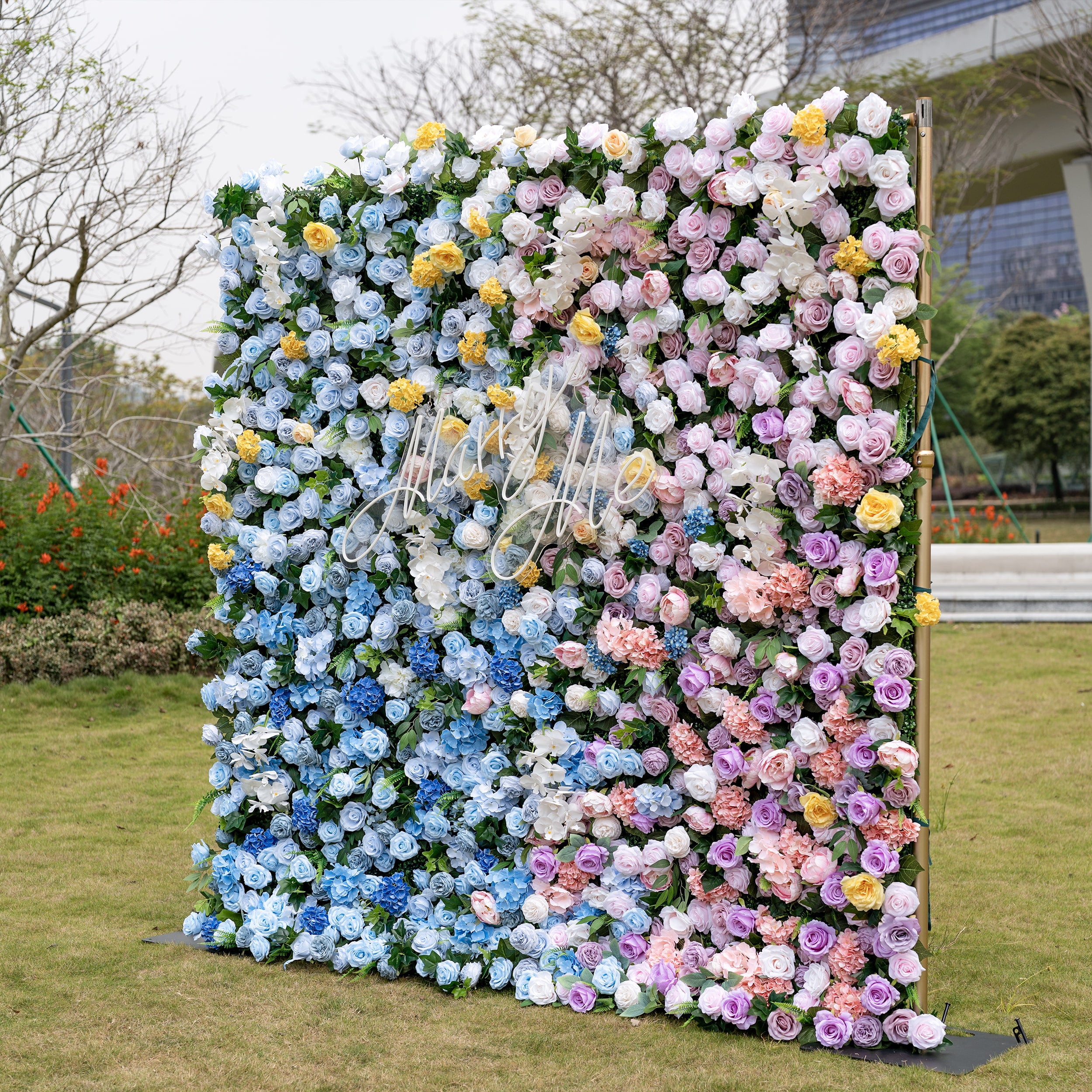 This is a stunning gradient artificial flower wall backdrop, set outdoors on a grassy lawn with an urban skyline in the background. It features a lush, dense arrangement of lifelike blooms: soft sky-blue delphiniums at the bottom, transitioning upward to creamy white roses, sunny yellow poppies, blush pink peonies, and lavender roses. This premium, reusable floral display creates an elegant, romantic focal point ideal for weddings, photo booths, bridal showers, and luxury event decor.