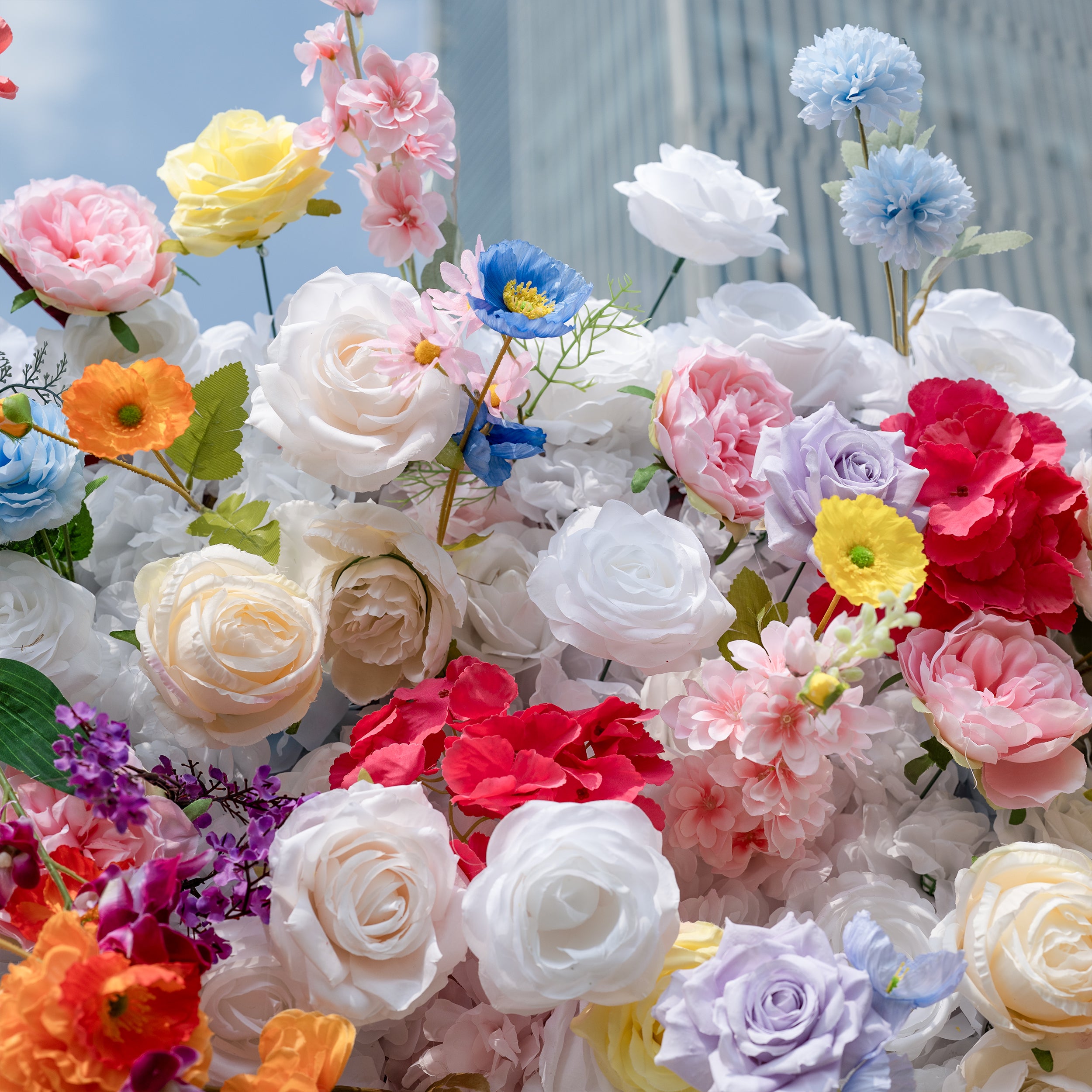 This is a lush, vibrant close-up of a premium artificial mixed-flower arrangement, brimming with lifelike blooms in a dreamy pastel and bright palette. It features creamy white roses, soft pink peonies, sunny yellow roses, vivid orange poppies, sky-blue delphiniums, deep red hydrangeas, lavender roses, and delicate filler flowers, set against a bright blue sky and modern building backdrop. The layered, realistic petals create a romantic, whimsical aesthetic, perfect for wedding backdrops, event decor, and p