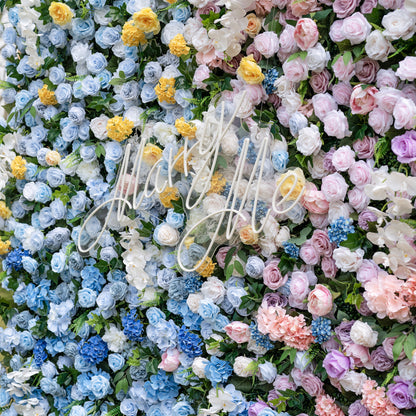 This is a stunning gradient artificial flower wall backdrop, centered with an elegant "Marry Me" neon script sign. It features a lush, dense arrangement of lifelike blooms: soft sky-blue delphiniums at the bottom, transitioning upward to creamy white roses, sunny yellow poppies, blush pink peonies, and lavender roses. This premium, reusable floral display creates a romantic, showstopping focal point ideal for wedding proposals, ceremonies, photo booths, and luxury event decor.