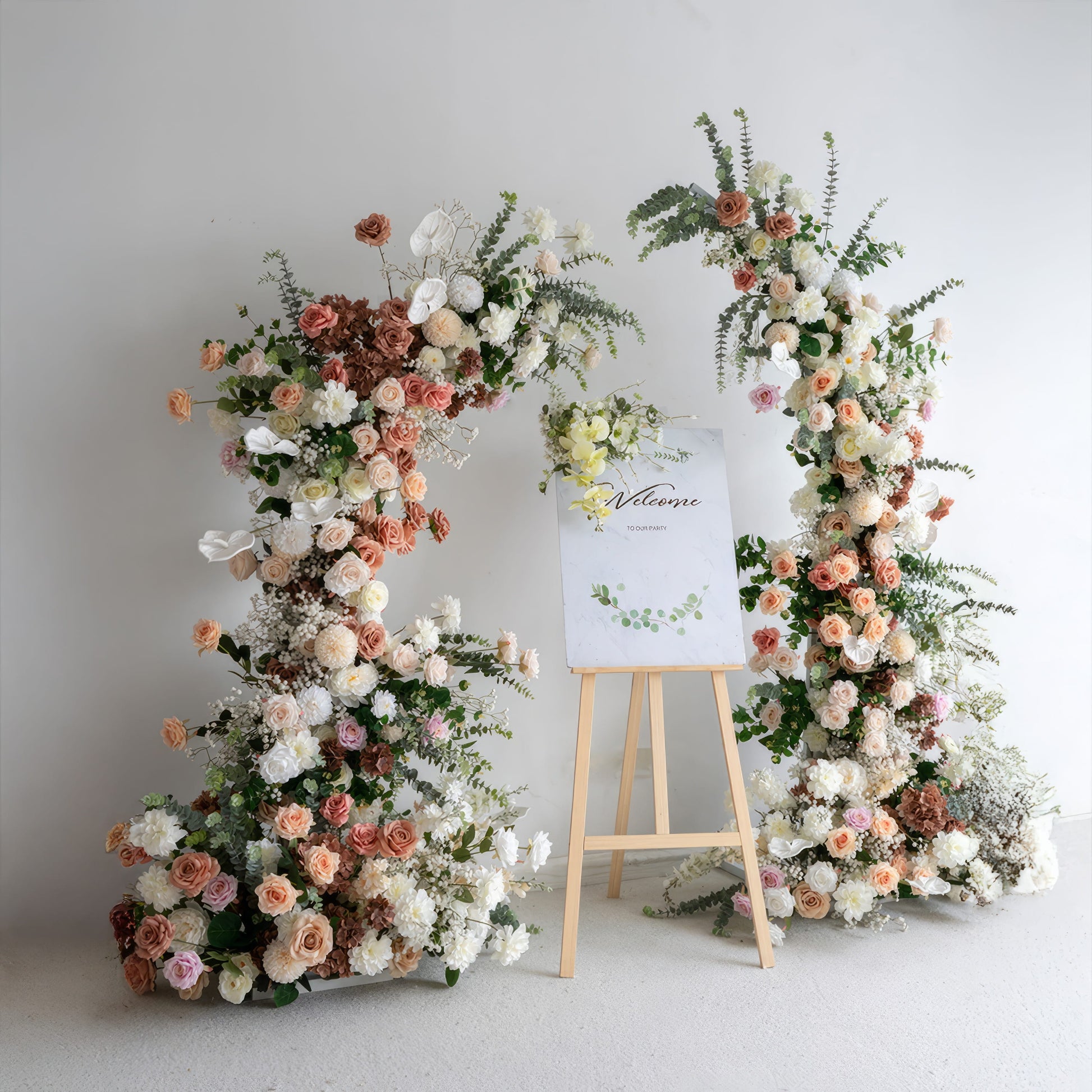 A romantic, asymmetrical floral arch from Valar Flowers, crafted with a soft, warm palette of blush pink, terracotta, and creamy white blooms—including roses, peonies, and delicate baby's breath—intertwined with silvery eucalyptus and lush greenery. A "Welcome" sign on a wooden easel rests between the arch, creating a dreamy, elegant, and sophisticated aesthetic ideal for luxury weddings and editorial photoshoots.
