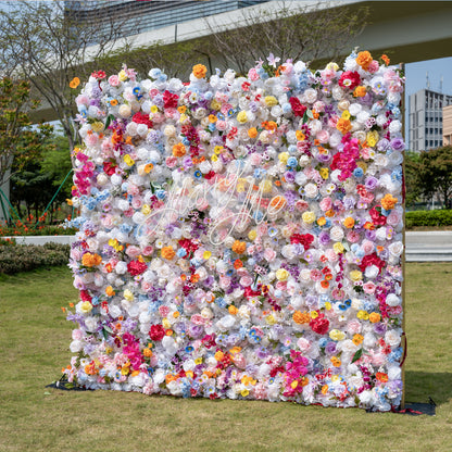 This is a showstopping, dense artificial flower wall backdrop from Valar Flowers, bursting with a vibrant, rainbow-hued mix of lifelike blooms. Set outdoors on a grassy lawn with an urban skyline in the background, it features creamy white roses, soft pink peonies, sunny yellow poppies, vivid orange carnations, sky-blue delphiniums, deep magenta orchids, and delicate filler flowers, creating a lush, romantic focal point perfect for weddings, photo booths, and luxury event decor.