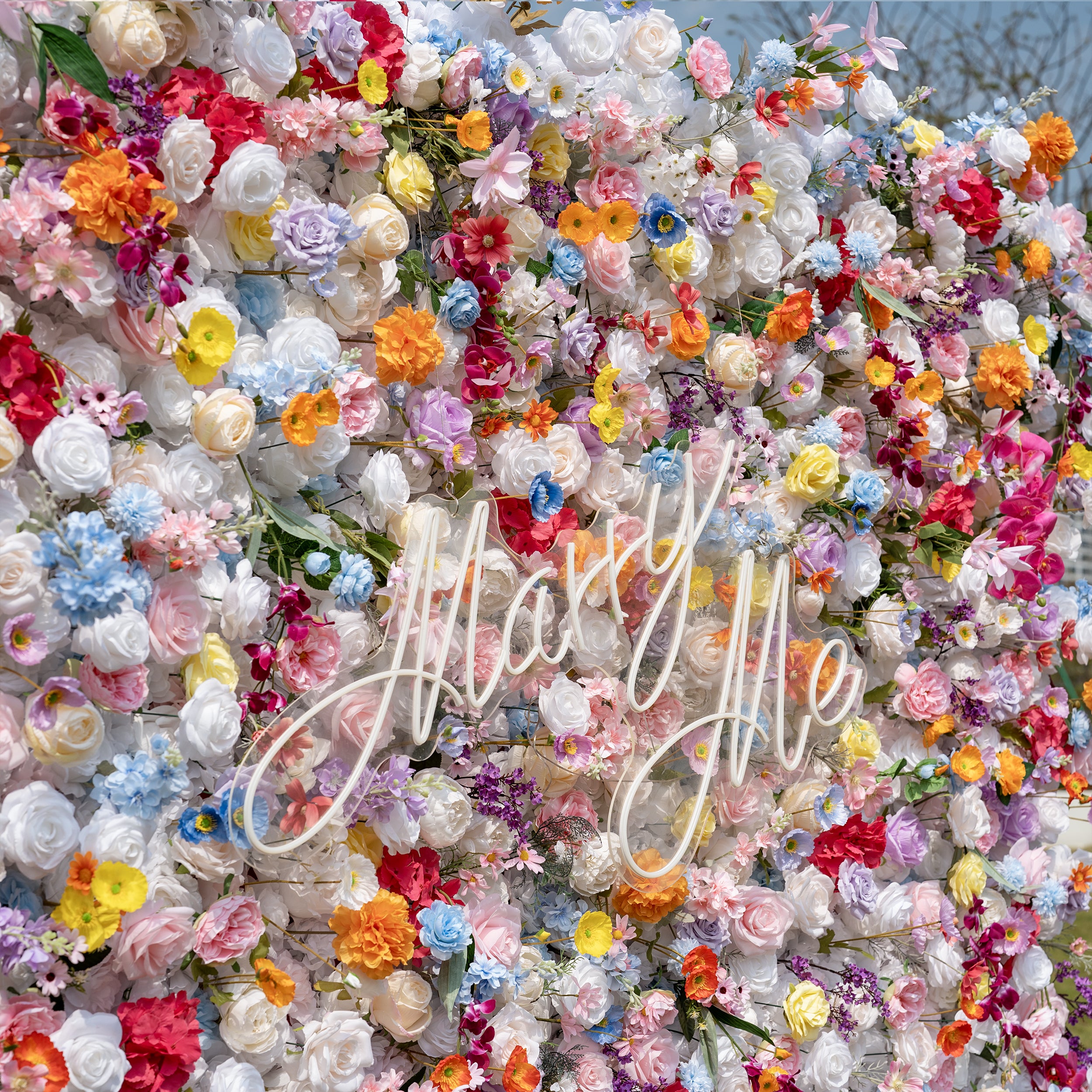 This is a breathtaking, dense artificial flower wall backdrop featuring the iconic "Just Married" script sign at its center. Bursting with a vibrant, rainbow spectrum of lifelike blooms—creamy white roses, soft pink peonies, sunny yellow poppies, vivid orange carnations, sky-blue delphiniums, magenta hydrangeas, and lavender orchids—this lush arrangement creates a romantic, showstopping focal point perfect for wedding ceremonies, reception decor, and photo booth styling.