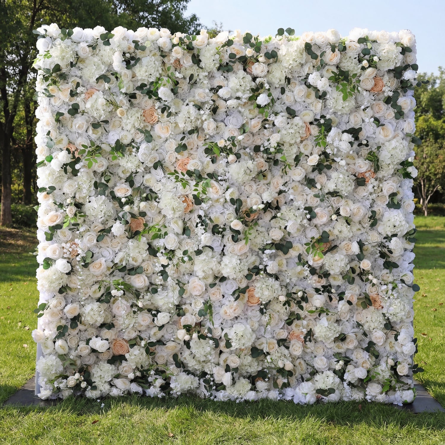 This opulent floral backdrop blends dense white blooms—roses, hydrangeas—with soft peach accents and delicate green foliage. Varied petal textures and subtle tonal gradients (ivory to warm peach) add depth, while natural sunlight enhances the lush, romantic appearance. The elegant neutral palette merges classic sophistication with organic fullness, making it a refined focal point ideal for weddings, formal events, or upscale outdoor celebrations.
