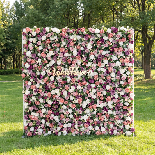 Vibrant mixed-tone artificial flower wall mounted on a metal stand outdoors on lush green grass, layered with blush peach, crisp white, dusty purple roses and fresh green foliage. Centered elegant golden "Valar Flowers" custom signage adds brand appeal. This reusable zero-maintenance floral backdrop suits wedding photo booths, bridal events, boutique displays and multi-pastel social media shoots for rich, whimsical romantic decor.