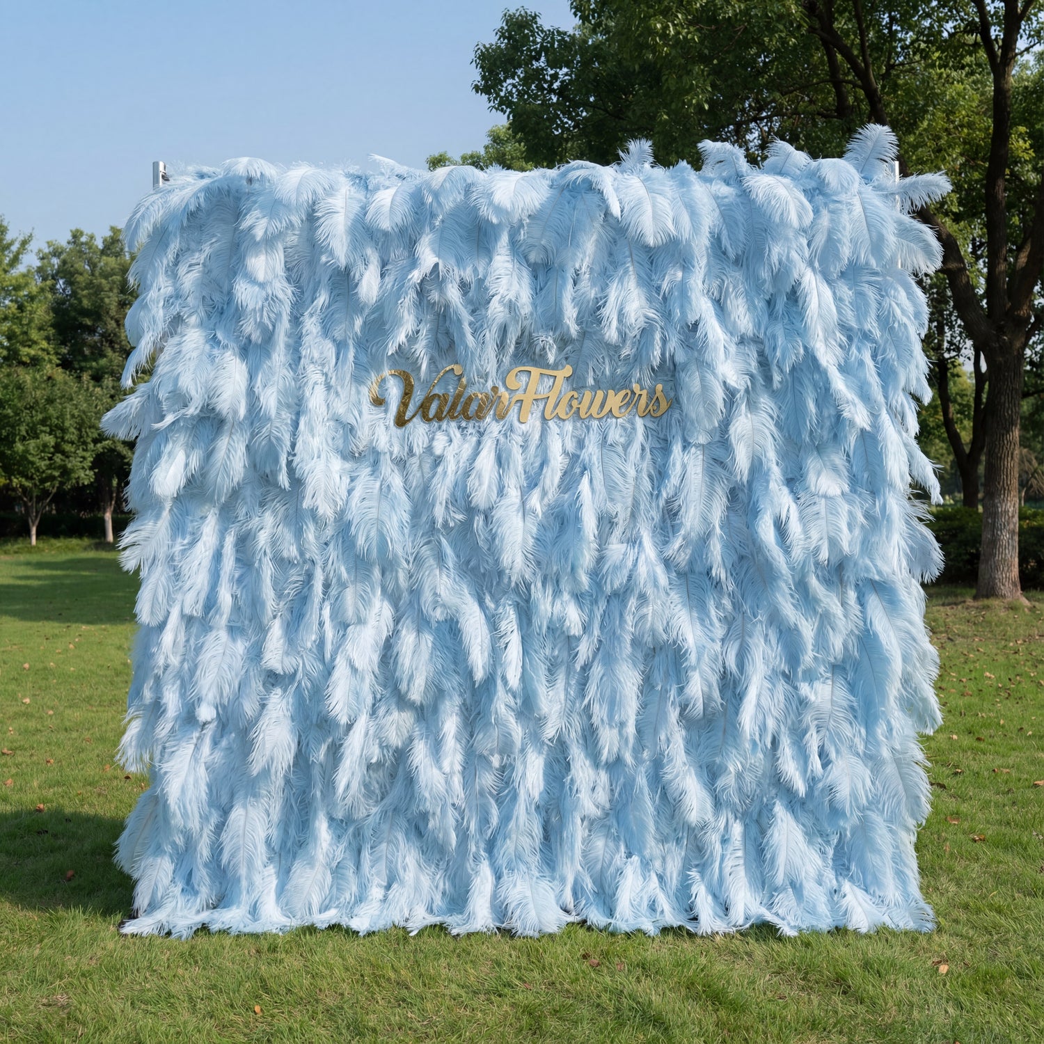 This luxurious backdrop (set on sunlit grass, tree/blue sky backdrop) is draped in soft, layered light blue ostrich feathers, with subtle gradients from pale sky-blue to deeper powder-blue. A polished gold “ValarFlowers” sign anchors the center, glowing against the airy, plush texture. Natural sunlight enhances the feathery depth, merging dreamy pastels with opulent metallic warmth to create an elegant, whimsical focal point for upscale outdoor events.