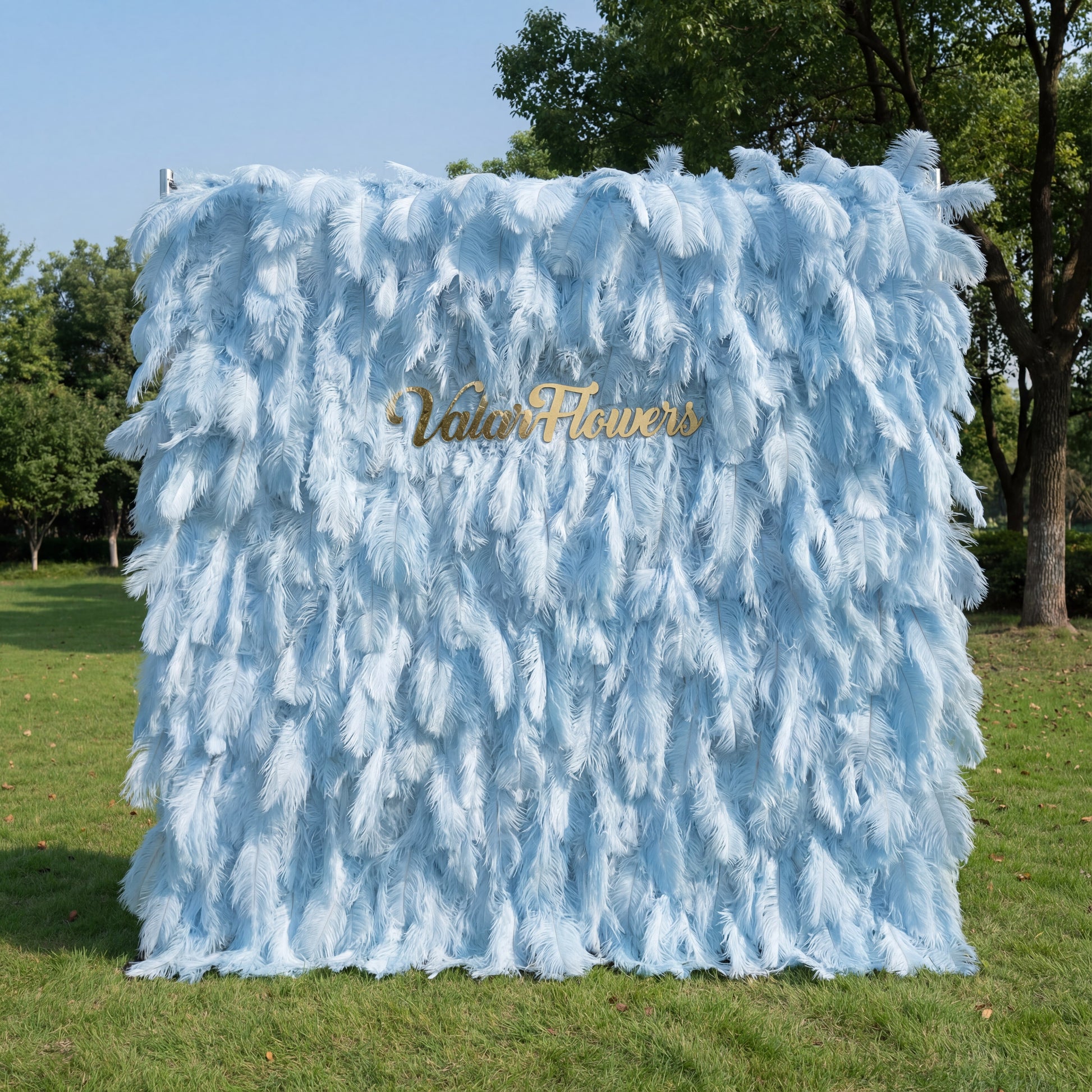 This luxurious backdrop (set on sunlit grass, tree/blue sky backdrop) is draped in soft, layered light blue artificial feathers, with subtle gradients from pale sky-blue to deeper powder-blue. A polished gold “ValarFlowers” sign anchors the center, glowing against the airy, plush texture. Natural sunlight enhances the feathery depth, merging dreamy pastels with opulent metallic warmth to create an elegant, whimsical focal point for upscale outdoor events.