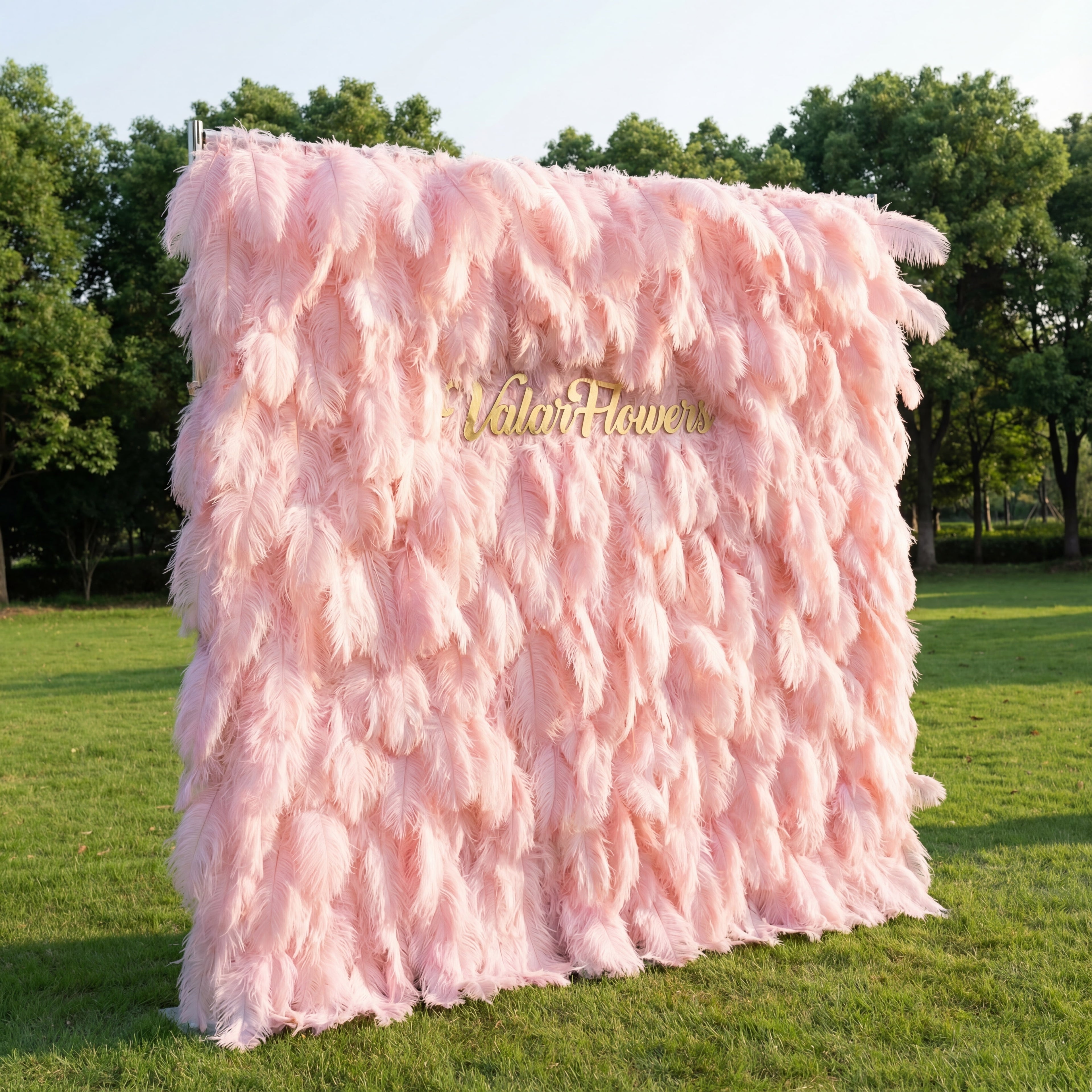 This whimsical backdrop (set on sunlit grass, tree backdrop) is draped in lush, layered blush-pink ostrich feathers, with soft gradients from pale rose to deeper peony. A polished gold “ValarFlowers” sign glows against the plush, airy plumage. Natural sunlight enhances the feathery texture’s depth, merging dreamy pastel softness with opulent metallic warmth to create a romantic, elegant focal point—ideal for upscale outdoor events or delicate celebrations.