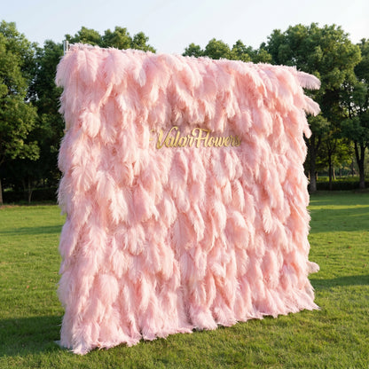 This whimsical backdrop (set on sunlit grass, tree backdrop) is draped in lush, layered blush-pink ostrich feathers, with soft gradients from pale rose to deeper peony. A polished gold “ValarFlowers” sign glows against the plush, airy plumage. Natural sunlight enhances the feathery texture’s depth, merging dreamy pastel softness with opulent metallic warmth to create a romantic, elegant focal point—ideal for upscale outdoor events or delicate celebrations.