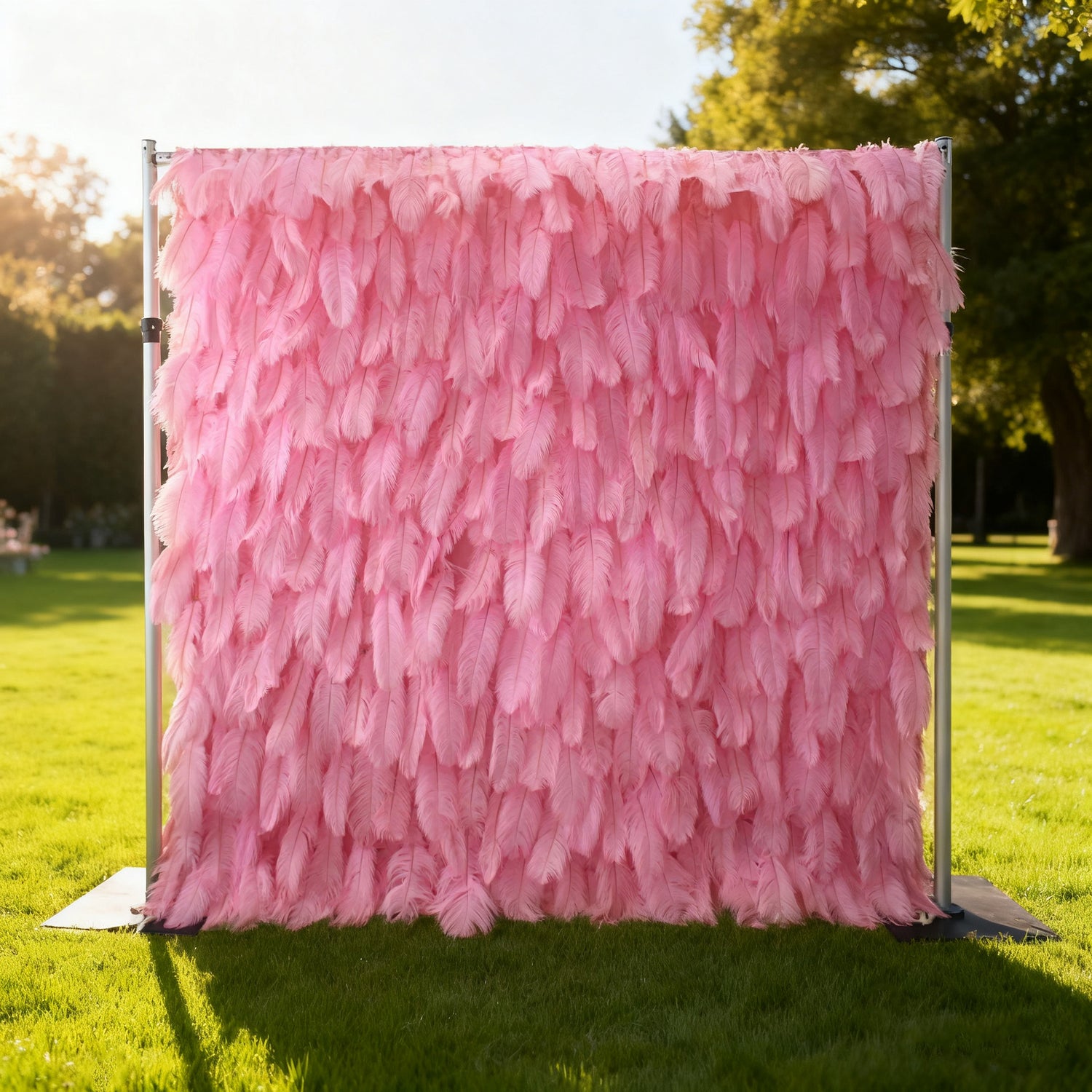 This image features a beautiful pink feather backdrop by Valar Flowers, set outdoors on a lush green lawn. The backdrop, composed of densely arranged soft pink feathers, creates a dreamy and elegant display. Supported by a sleek metal frame, it exudes a sense of whimsy and sophistication, perfect for adding a touch of charm to events like weddings or photo shoots, with sunlight and trees enhancing its enchanting appeal.