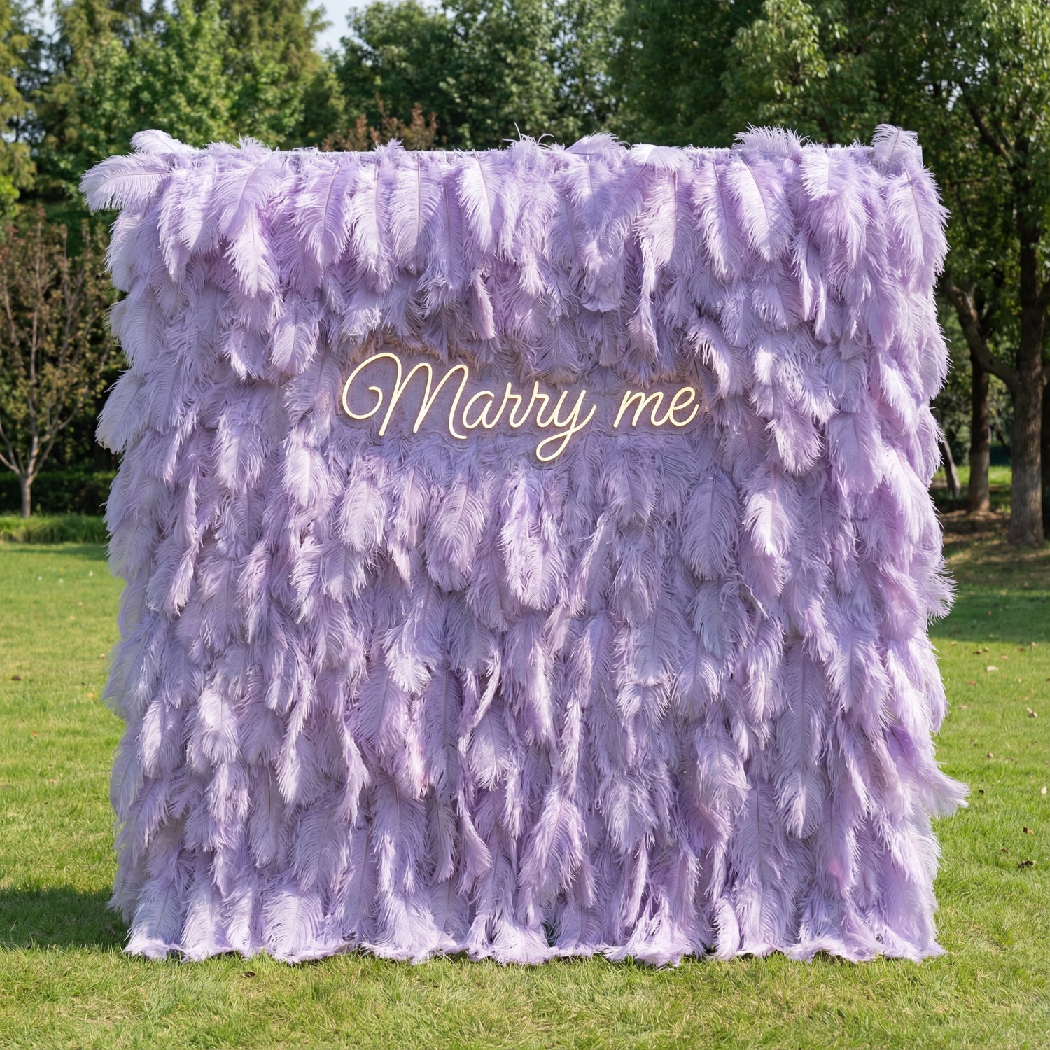 This ethereal backdrop (set on sunlit grass, tree backdrop) is draped in soft, layered lavender ostrich feathers, creating a plush, dreamy texture with subtle pale-to-deep purple gradients. A glowing cursive “Marry me” sign nestles at the center, casting a gentle light against the feathery surface. Natural sunlight enhances its airy, romantic vibe, merging opulent whimsy with heartfelt charm to serve as a striking, intimate focal point for proposals or upscale outdoor celebrations.