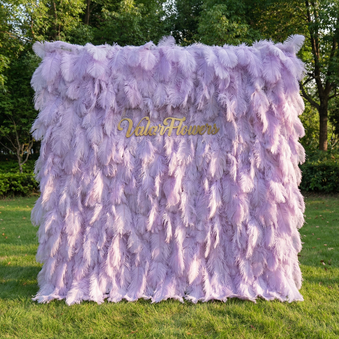 This luxurious backdrop (set on sunlit grass, tree backdrop) is covered in soft, layered lavender ostrich feathers, creating a plush, ethereal texture with subtle pale-to-deep purple gradients. A polished gold “ValarFlowers” sign anchors the center, glowing against the dreamy, muted palette. Natural light enhances the feathery, airy vibe, merging opulent elegance with whimsical charm to serve as a striking focal point for upscale events or romantic photo settings.