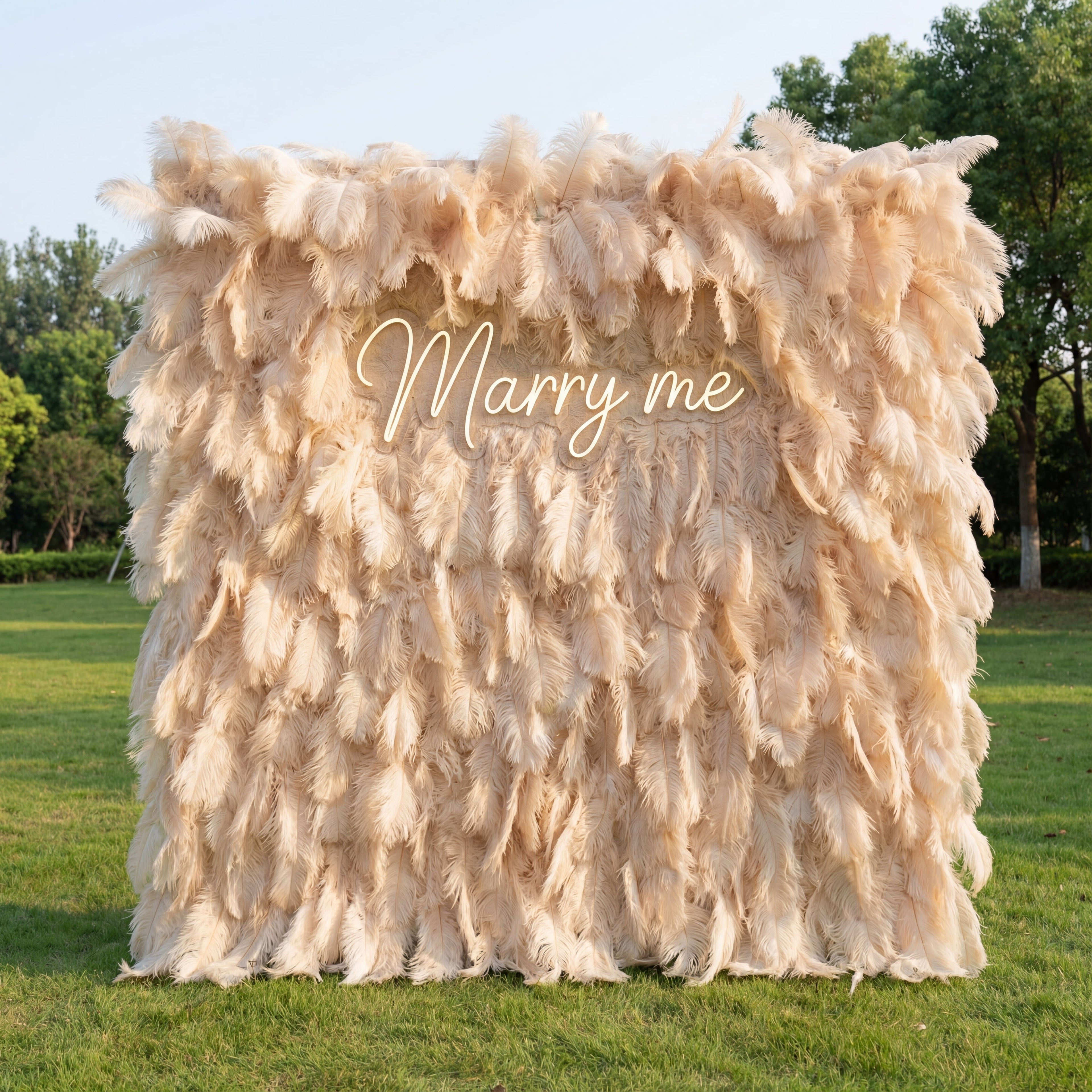 This romantic backdrop is draped in dense, fluffy beige ostrich feathers, creating a soft, opulent texture with subtle pale peach gradients. A glowing cursive “Marry me” sign nestles at the center, its warm light complementing the plush plumage. Natural sunlight highlights the feathers’ wispy, delicate surfaces, merging glamorous elegance with heartfelt warmth to form an ethereal focal point—perfect for intimate proposals or dreamy outdoor celebrations.
