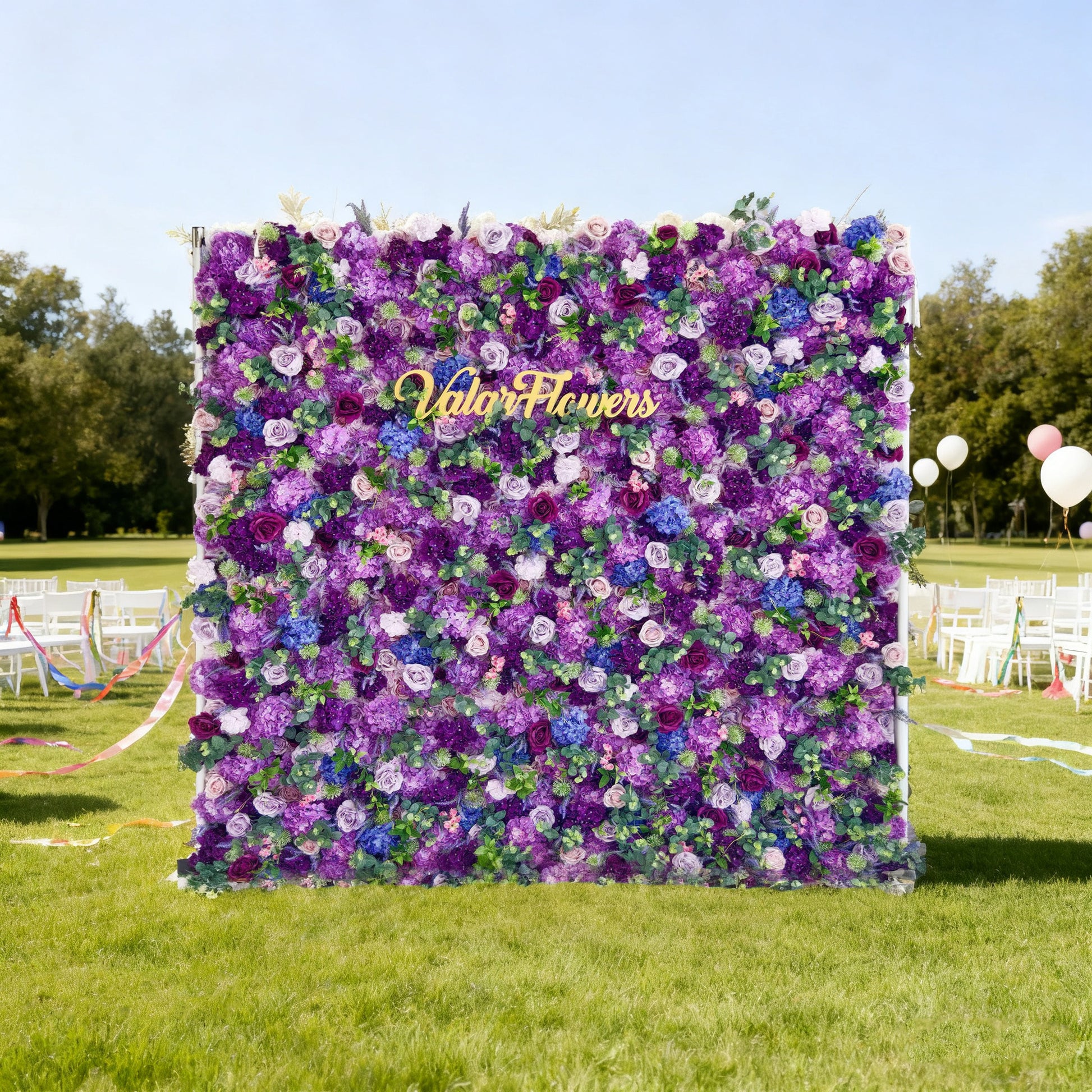 This image features a stunning floral wall by Valar Flowers, showcasing a vibrant mix of purple, lavender, and blue blooms. The golden "Valar Flowers" logo stands out prominently. Set outdoors on a lush green lawn, with white chairs, colorful ribbons, and balloons in the background, it creates a festive and elegant atmosphere, perfect for events like weddings or parties, highlighting the brand's beautiful floral craftsmanship.
