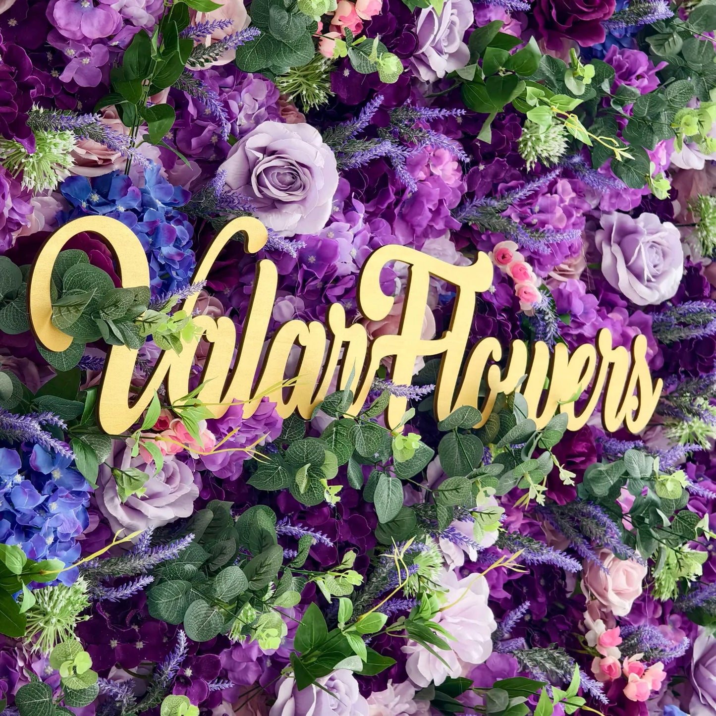 This image showcases a close - up of a lush floral wall by Valar Flowers. The arrangement is a vibrant mix of purple, blue, and lavender flowers, including roses and hydrangeas, interspersed with green foliage. The golden "Valar Flowers" text stands out prominently against the richly colored blooms, creating a visually striking and elegant display, perfect for events or as a decorative backdrop. 