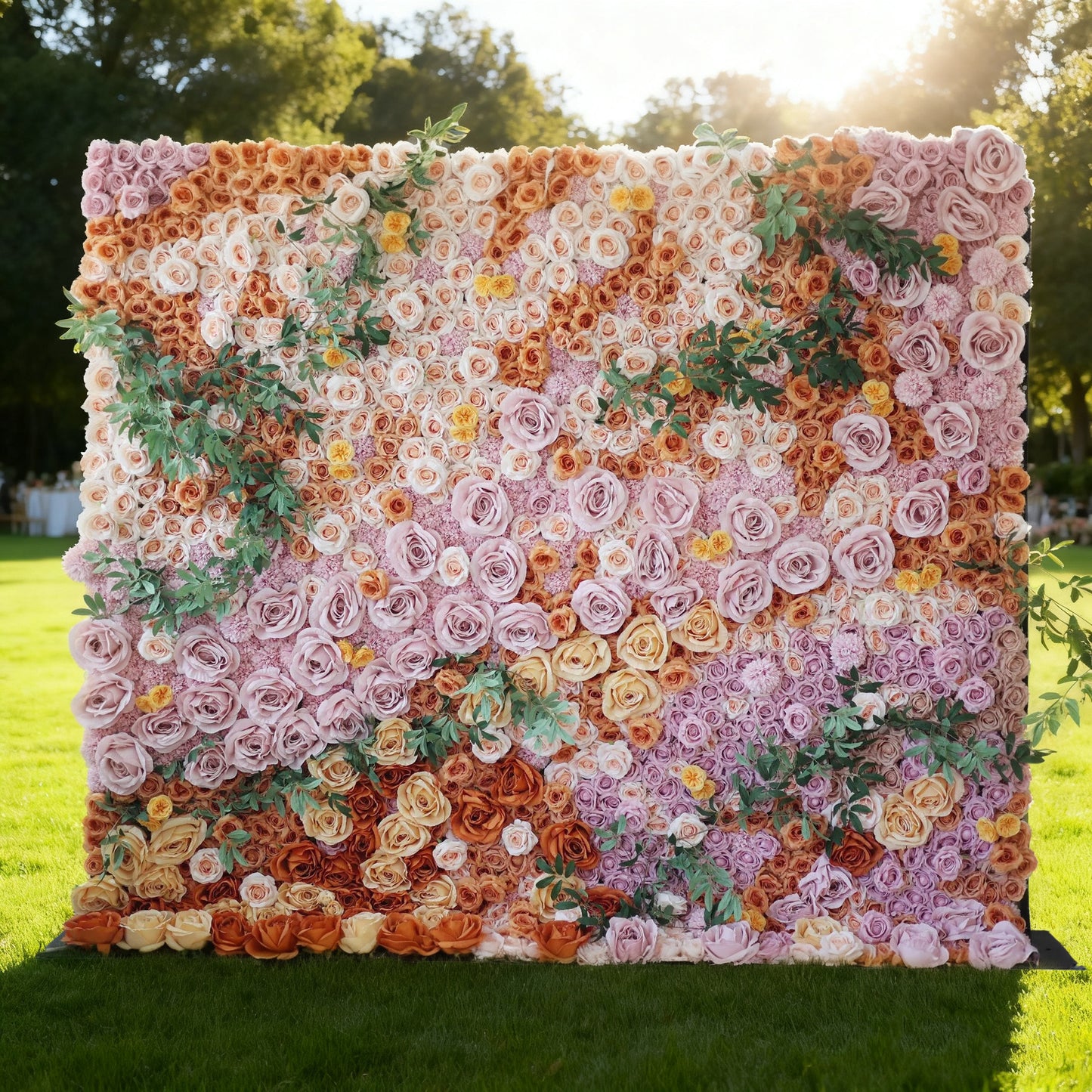 This image showcases a stunning floral wall by Valar Flowers, set outdoors on a lush green lawn. It's densely covered with artificial flowers in a rich palette of colors, including shades of pink, orange, and yellow, along with green foliage. Bathed in sunlight with trees in the background, the floral wall exudes a vibrant and elegant atmosphere, perfect for decorative use in events like weddings or photo shoots.