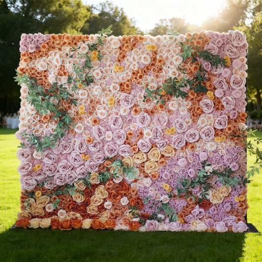 This image showcases a stunning floral wall by Valar Flowers, set outdoors on a lush green lawn. It's densely covered with artificial flowers in a rich palette of colors, including shades of pink, orange, and yellow, along with green foliage. Bathed in sunlight with trees in the background, the floral wall exudes a vibrant and elegant atmosphere, perfect for decorative use in events like weddings or photo shoots.