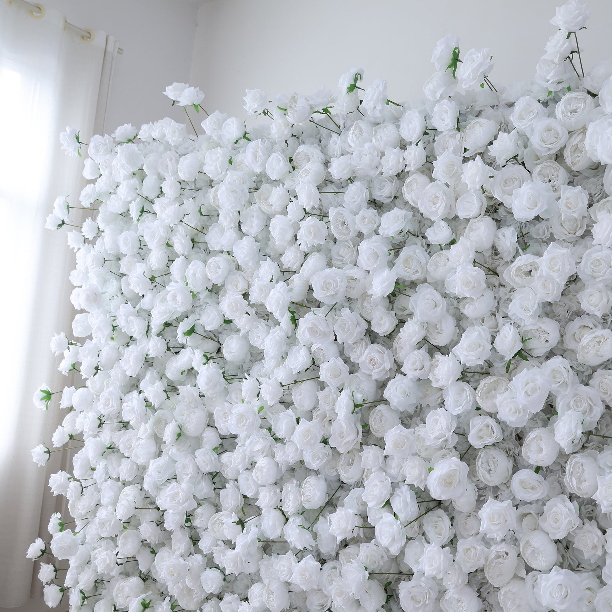 This image showcases a dense arrangement of white artificial roses, forming a lush and voluminous floral display. The flowers are tightly packed, with their soft, creamy petals and subtle green stems creating a sense of purity and elegance. Set indoors, likely near a window with light filtering through, the scene exudes a serene and sophisticated atmosphere. It's ideal for decorative purposes in events or as a striking interior backdrop, evoking feelings of natural beauty and refinement.