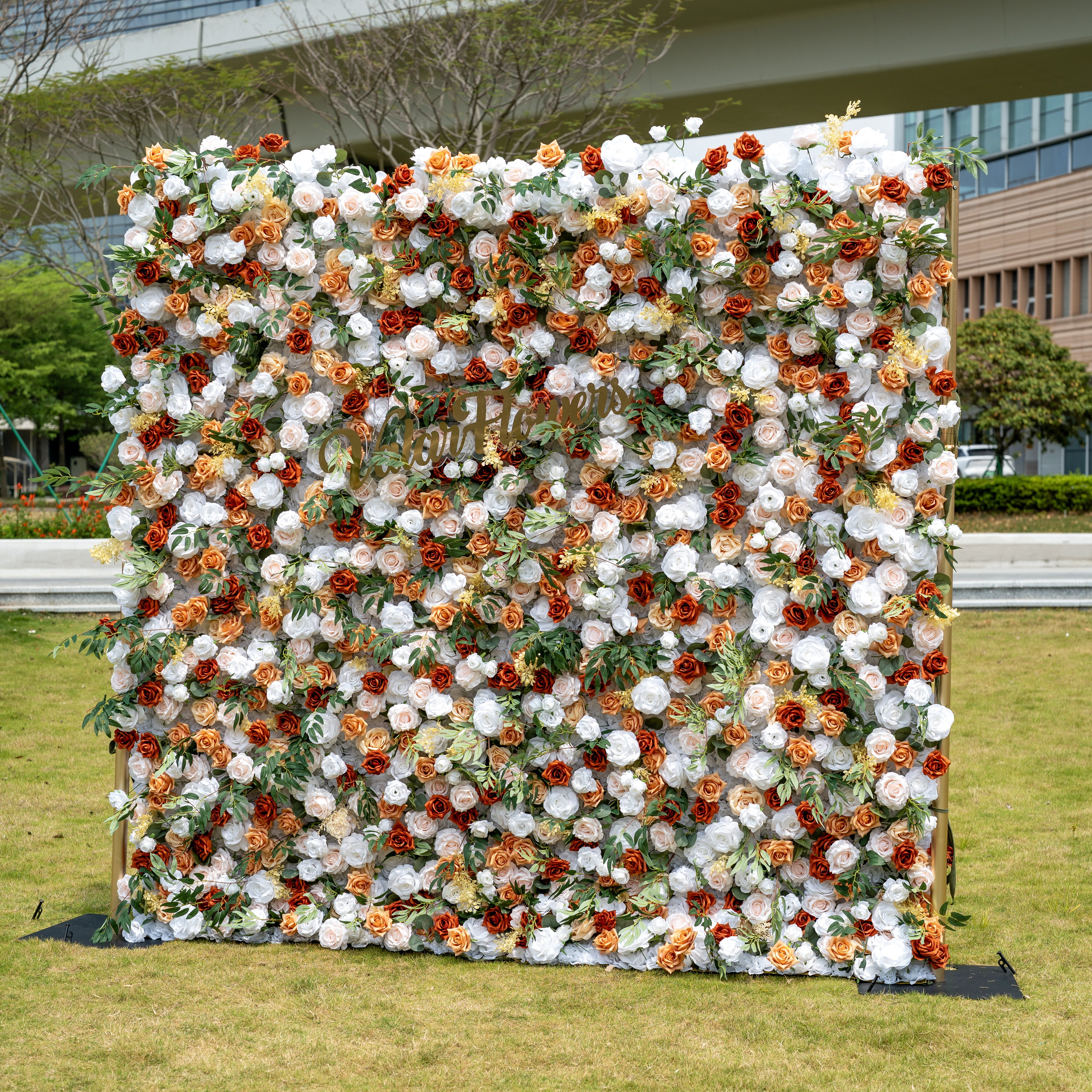 This is a stunning, full-size artificial flower wall backdrop, set outdoors on a grassy lawn with an urban skyline in the background. It features a lush, dense arrangement of lifelike blooms in a warm terracotta and cream palette: burnt orange roses, creamy white peonies, soft peach roses, and vibrant green foliage. This premium, reusable floral display creates a timeless, romantic focal point, perfect for rustic weddings, fall events, photo booths, and luxury outdoor decor.