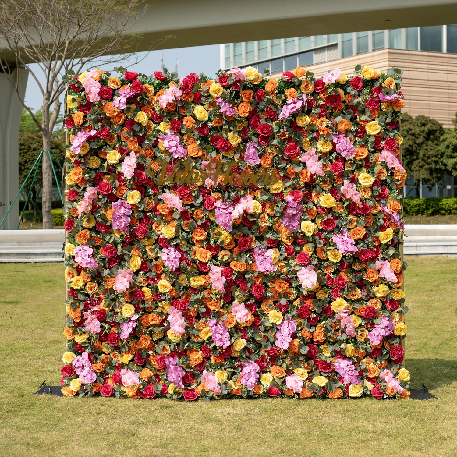 This is a full-size 8x8ft ValarFlowers premium multicolor artificial flower wall backdrop, featuring a lush, dense arrangement of lifelike burgundy, orange, yellow roses, pink hydrangeas, and realistic eucalyptus greenery, centered with a custom "Valar Flowers" logo sign. This high-quality, reusable floral display creates a warm, romantic focal point, ideal for wedding backdrops, photo booths, garden parties, and luxury outdoor event styling.