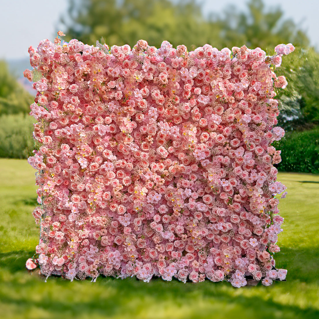 This photo features a large, square floral backdrop densely packed with soft pink roses, delicate pink orchids, and small pink flowers. Set on a grassy field with greenery in the background, it exudes a serene and romantic charm. Ideal for events like a "Pink Blossom Garden Party" or an intimate wedding, it creates a dreamy and picturesque atmosphere.