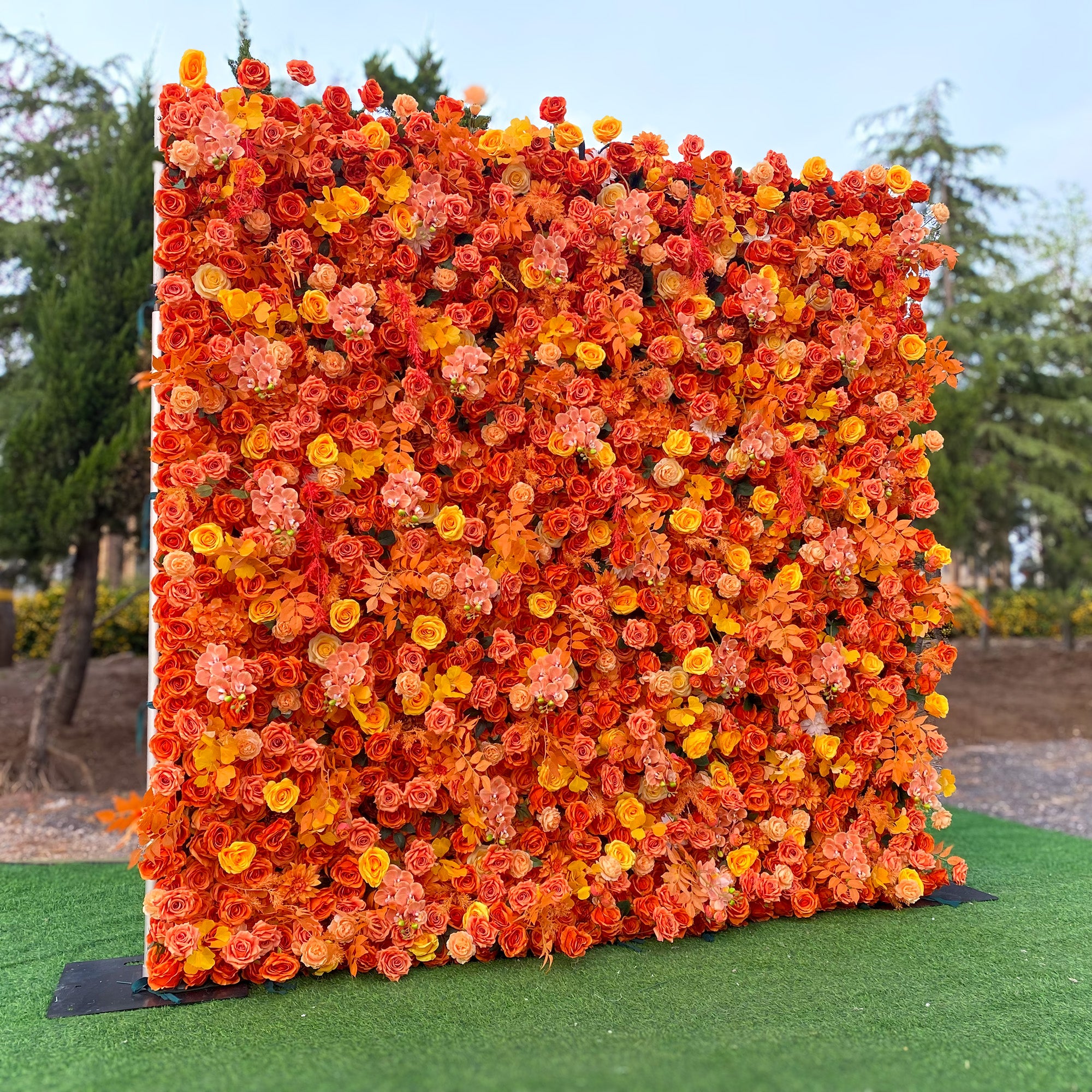 This image features a vibrant floral wall set outdoors on a green lawn, with trees in the background. The wall is densely covered with artificial flowers predominantly in shades of orange, ranging from bright tangerine to soft peach, along with touches of yellow. The flowers appear to be mainly roses and some small, clustered blooms. The overall style is bold and warm, creating a striking and eye - catching display. 