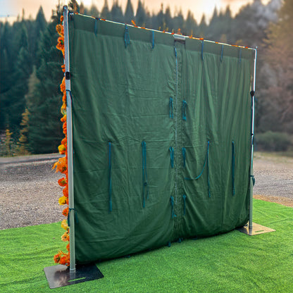 This image depicts a setup featuring a green fabric backdrop supported by a metal frame with square bases. The frame is adorned on one side with a vibrant arrangement of artificial flowers in shades of orange and yellow, likely roses and other assorted blooms. The style is functional with a touch of decorative flair, suitable for creating a partition or backdrop for events. 