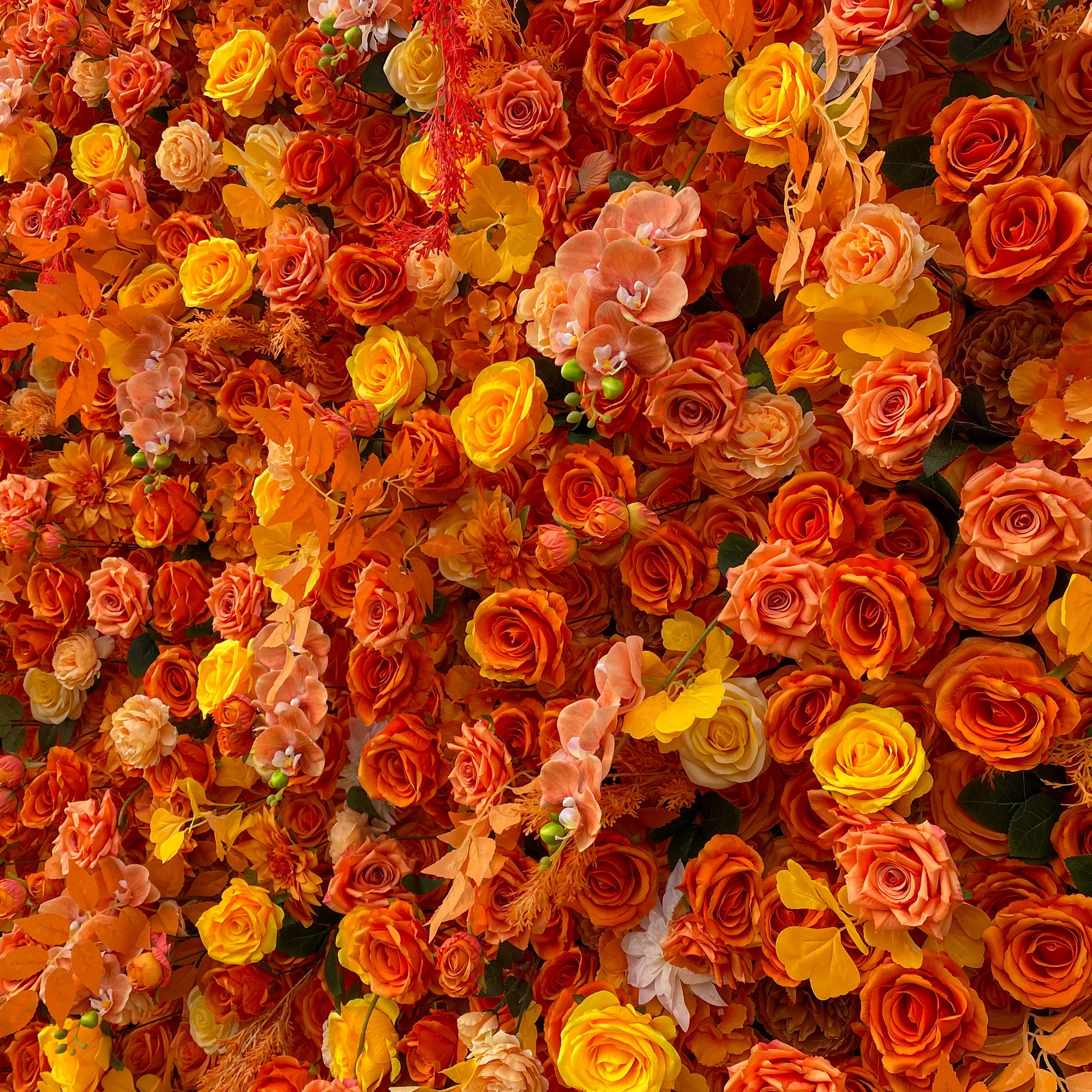 This image showcases a lush floral arrangement predominantly featuring artificial roses in a rich palette of warm colors, including vibrant orange, sunny yellow, and soft peach. Interspersed among the roses are delicate orchids and other assorted blooms, along with some foliage elements. The overall style is dense and vibrant, creating a warm and inviting autumnal - inspired visual display. 
