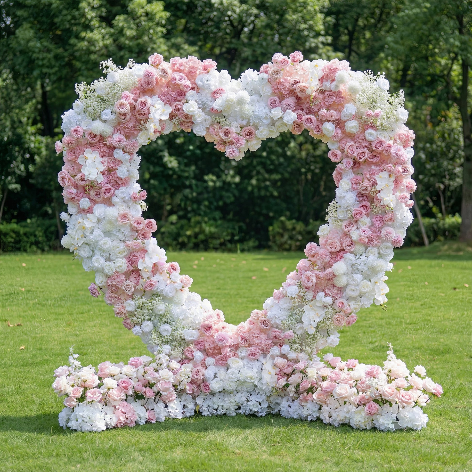 This romantic outdoor heart-shaped floral arch is crafted from a lush blend of blush pink, deep rose, and crisp white blooms, including roses, hydrangeas, and delicate baby's breath. The dense, layered design creates a dreamy, three-dimensional texture, while a matching floral arrangement at its base completes the look. Set on a lush green lawn framed by leafy trees, it exudes timeless romance, making it a stunning focal point for weddings, engagement photoshoots, or romantic celebrations.