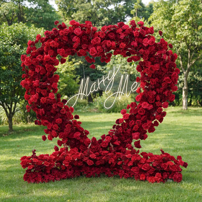 This outdoor setup features a lush, heart-shaped arch dense with deep red roses, resting on green grass framed by trees. A glowing "Marry Me" neon sign (cursive, light-lit) is centered within the blooms, its soft glow contrasting with the rich, passionate hues. The bold, romantic display exudes warmth, perfect for proposal setups, wedding decor, or intimate romantic events.