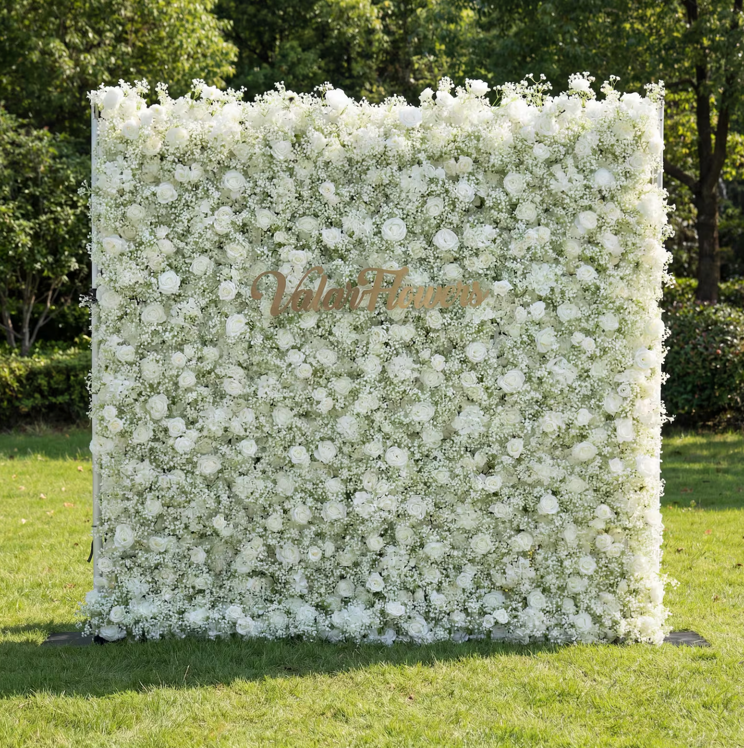 This is a lush, all-white artificial flower wall backdrop from **Valar Flowers**, densely packed with white roses and delicate baby's breath. A gold "ValarFlowers" sign overlays the center, set against a green lawn and trees, creating a clean, elegant, and romantic aesthetic ideal for weddings, photo shoots, or special events.