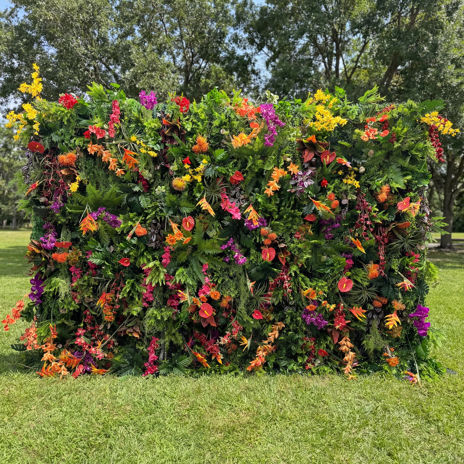 This Valar Flowers tropical flower wall backdrop is a lush, vibrant tapestry of exotic blooms and foliage, featuring fiery orange heliconias, bright red anthuriums, deep purple orchids, sunny yellow sprays, and rich green ferns. Set against a sunlit lawn, it creates a bold, jungle-inspired focal point ideal for outdoor weddings, summer parties, and lively photo opportunities, blending natural vibrancy with eye-catching color.