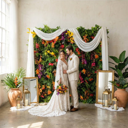 This Valar Flowers tropical flower wall backdrop is a lush, vibrant tapestry of exotic blooms and foliage, featuring fiery orange heliconias, bright red anthuriums, deep purple orchids, sunny yellow sprays, and rich green ferns, framed by flowing sheer white drapes. Paired with elegant gold mirrors, terracotta planters, and lanterns, it creates a romantic, bohemian focal point ideal for indoor weddings, ceremonies, and timeless photo opportunities.