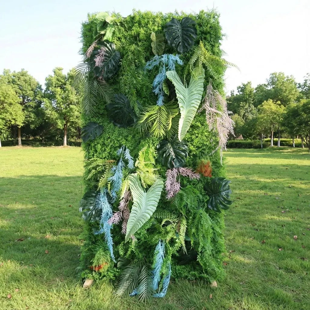 This vibrant greenery backdrop blends lush emerald foliage—monstera leaves, ferns, and palm fronds—with playful accents: pale blue feathery fronds and soft pink plumes, plus tiny rust accents. Varied textures (glossy, delicate, feathery) and tonal depth (from deep green to pastel) create a whimsical, exotic vibe. Natural sunlight highlights the lifelike surfaces, making it a striking, eclectic focal point ideal for creative outdoor events or tropical-chic celebrations.