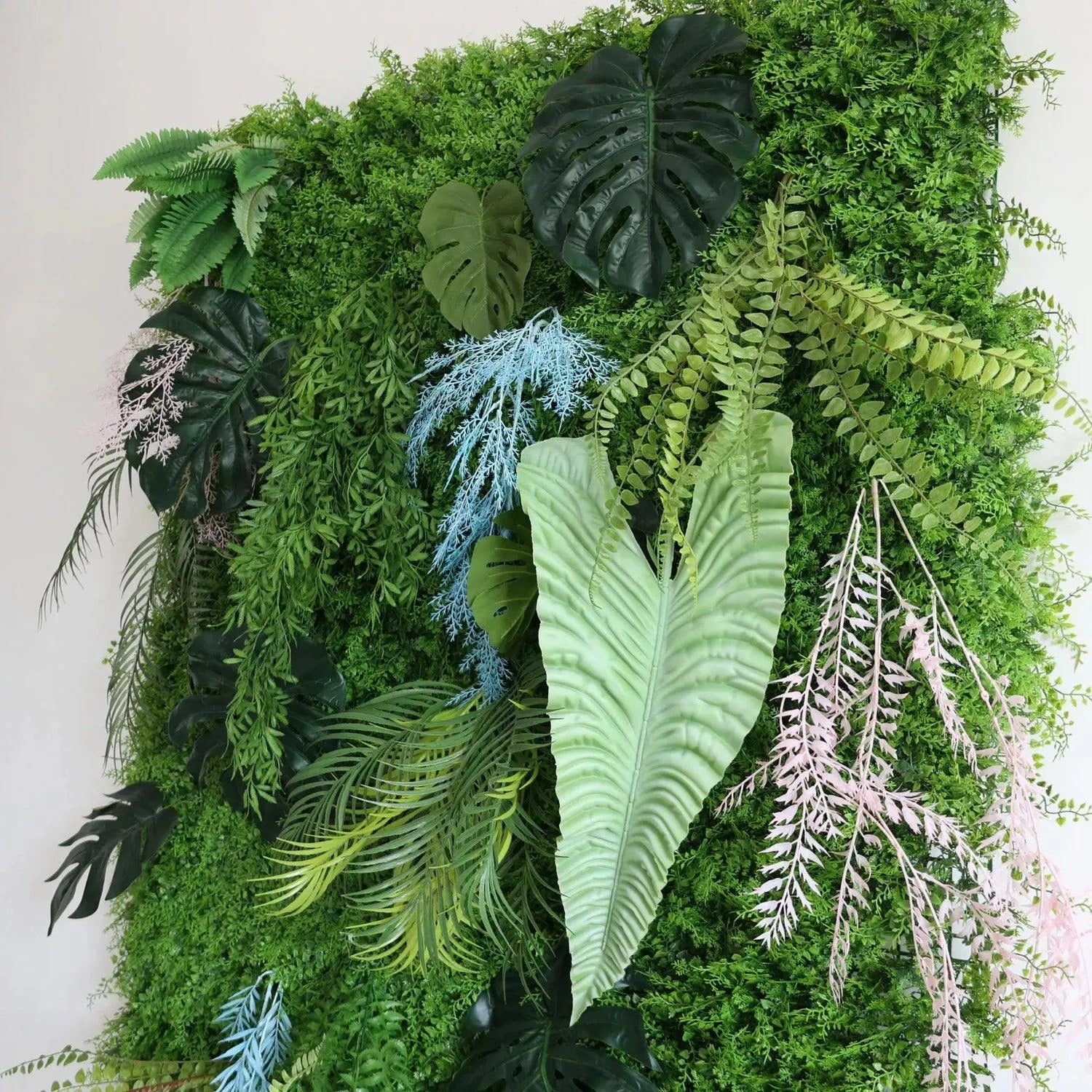 This photo showcases a lush artificial plant wall. It features prominent Monstera deliciosa leaves with their iconic splits, various fern types with delicate fronds, and palm-like leaves. Also, there are unique light blue and pinkish-lavender artificial plants, plus some orange-tinged elements. The dense greenery creates a vibrant, low-maintenance natural backdrop for interiors or events.