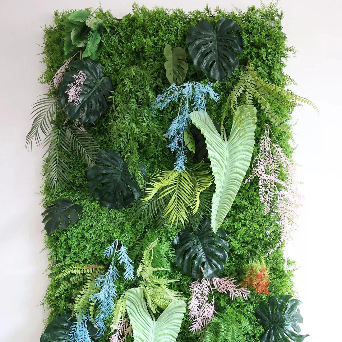 Palm-like leaves add a tropical touch. Among the greenery, non-green foliage offers unique color pops: light blue, pinkish-lavender artificial plants (likely ornamental grasses) weave through, contrasting the dominant green. Orange-tinged elements, mimicking flowering or autumnal plants, are also present. This vibrant, low-maintenance green wall serves as a natural backdrop, ideal for interiors or events.