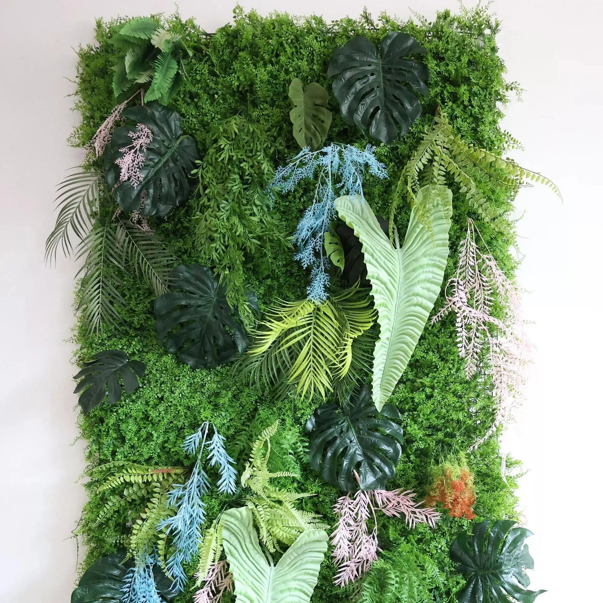 Palm-like leaves add a tropical touch. Among the greenery, non-green foliage offers unique color pops: light blue, pinkish-lavender artificial plants (likely ornamental grasses) weave through, contrasting the dominant green. Orange-tinged elements, mimicking flowering or autumnal plants, are also present. This vibrant, low-maintenance green wall serves as a natural backdrop, ideal for interiors or events.