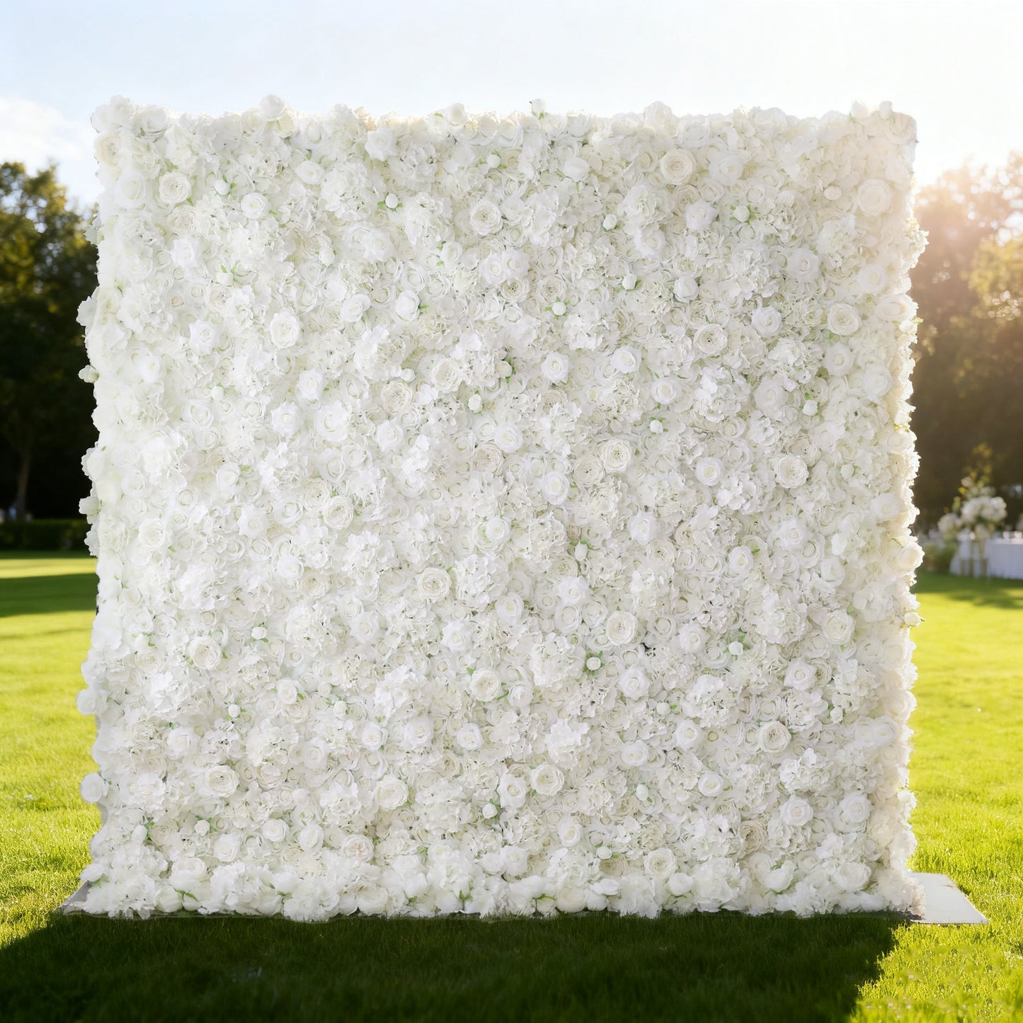 This image showcases a large, square floral wall outdoors on a lush green lawn. It's densely covered with pure white flowers, creating a clean, elegant, and serene display. Bathed in sunlight, with trees and a bright sky in the background, the scene exudes a sense of purity and natural beauty. From the brand 'Valar Flowers', this floral wall is perfect for decorative purposes in events or as a captivating backdrop.