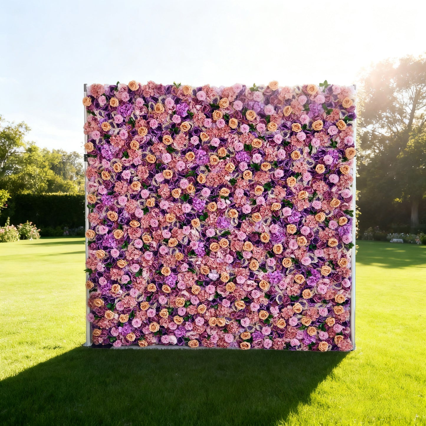 This image features a large, square floral wall set outdoors on a lush green lawn. It's densely covered with an array of flowers in shades of purple, pink, and peach. The vibrant blooms create a rich, colorful, and elegant display. Surrounded by trees and bathed in sunlight, the scene exudes a sense of natural beauty and charm, perfect for decorative purposes in events or as a picturesque backdrop.