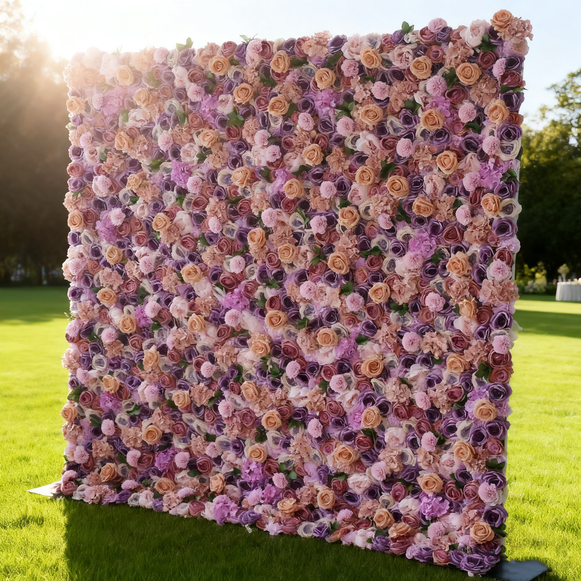 This image showcases a large, square floral wall set outdoors on a lush green lawn. It's densely covered with an array of flowers in shades of purple, pink, and peach. The vibrant blooms create a rich, colorful, and elegant display. Surrounded by trees and bathed in sunlight, the scene exudes a sense of natural beauty and charm, perfect for decorative purposes in events or as a picturesque backdrop.