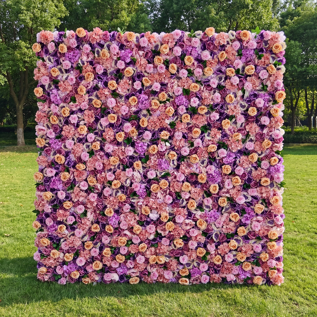 This vibrant outdoor floral backdrop features a dense, textured arrangement of artificial blooms in a romantic, sunset-inspired palette of blush, warm peach, and rich lavender. Lush roses and delicate filler flowers create a velvety, three-dimensional effect that exudes warmth and whimsy. Set on a sunlit green lawn framed by trees, it serves as a lively focal point for weddings, garden parties, or photoshoots seeking a bold, joyful aesthetic.
