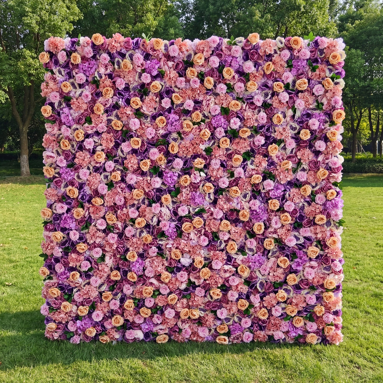 This vibrant outdoor floral backdrop features a dense, textured arrangement of artificial blooms in a romantic, sunset-inspired palette of blush, warm peach, and rich lavender. Lush roses and delicate filler flowers create a velvety, three-dimensional effect that exudes warmth and whimsy. Set on a sunlit green lawn framed by trees, it serves as a lively focal point for weddings, garden parties, or photoshoots seeking a bold, joyful aesthetic.