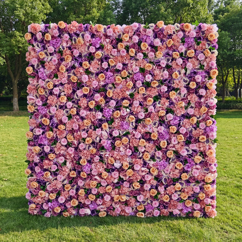This vibrant outdoor floral backdrop features a dense, textured arrangement of artificial blooms in a romantic, sunset-inspired palette of blush, warm peach, and rich lavender. Lush roses and delicate filler flowers create a velvety, three-dimensional effect that exudes warmth and whimsy. Set on a sunlit green lawn framed by trees, it serves as a lively focal point for weddings, garden parties, or photoshoots seeking a bold, joyful aesthetic.