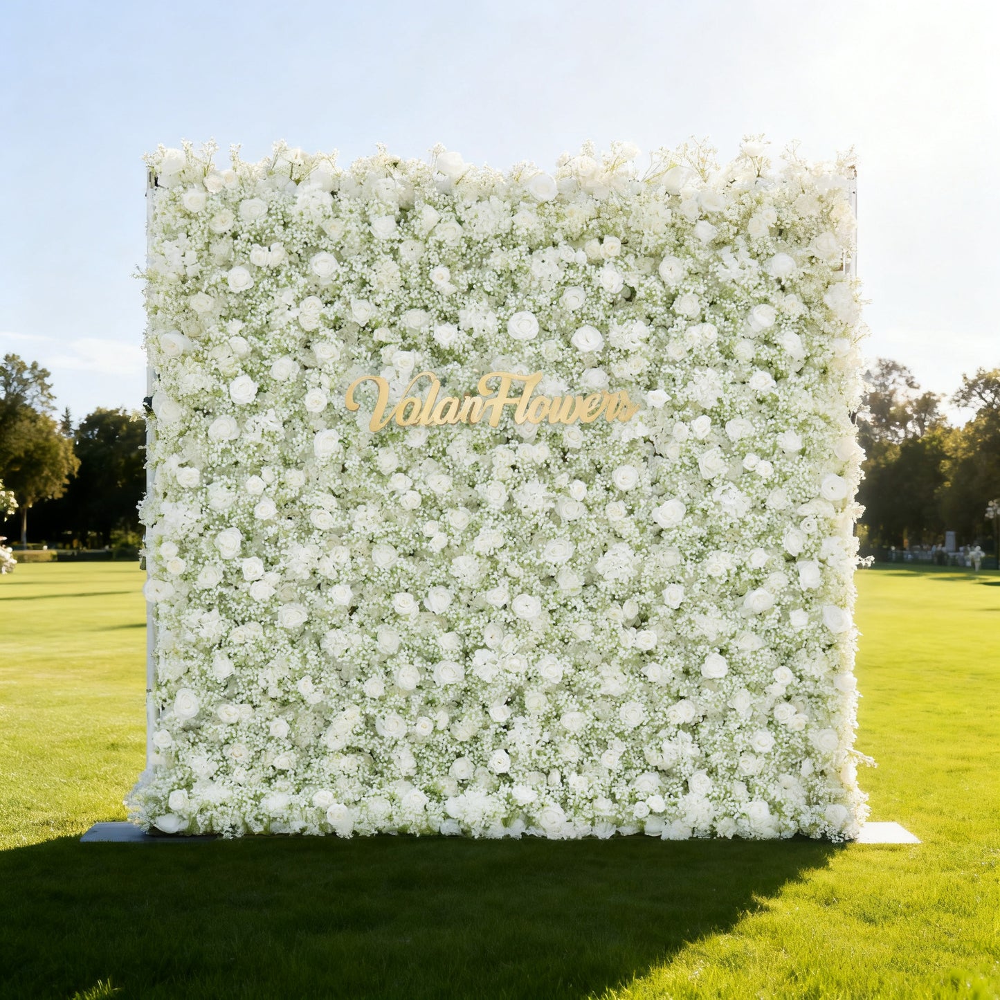 This image features a large, square floral wall densely covered with white flowers, including roses and baby's breath. "Valar Flowers" is displayed in elegant golden text. Set outdoors on a lush green lawn under a bright sky with trees in the background, it creates a pure, elegant, and serene atmosphere, perfect for events or decorative displays, exuding a sense of natural beauty and refinement.