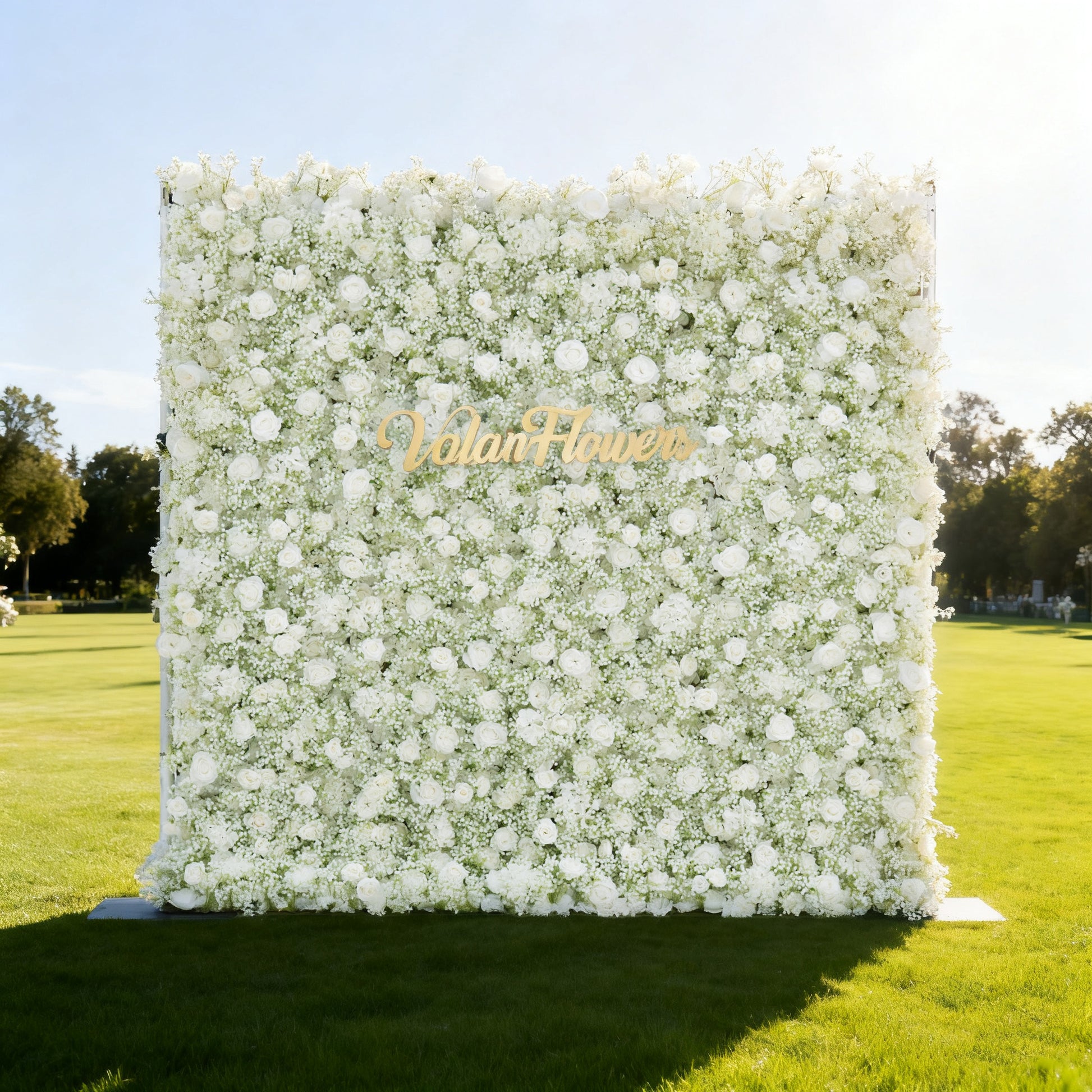 This image features a large, square floral wall densely covered with white flowers, including roses and baby's breath. "Valar Flowers" is displayed in elegant golden text. Set outdoors on a lush green lawn under a bright sky with trees in the background, it creates a pure, elegant, and serene atmosphere, perfect for events or decorative displays, exuding a sense of natural beauty and refinement.