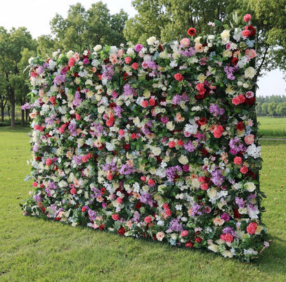 A sprawling outdoor floral wall by Valar Flowers stands on lush green grass, backed by tall trees. It bursts with a vibrant mix of bold red roses, soft pink peonies, deep purple stock, creamy white carnations, and peach gerberas, all nestled in dense green foliage. This eye-catching, full-bloom design creates a cheerful, festive atmosphere, perfect for garden weddings, outdoor receptions, or vibrant event photo backdrops.