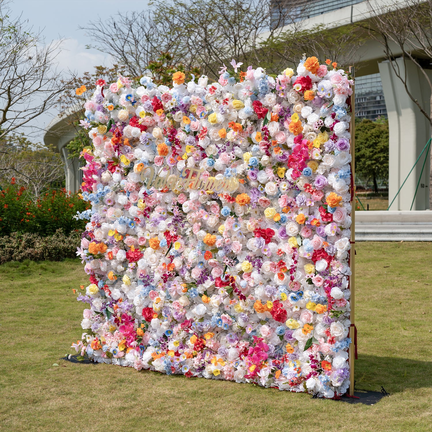 This is a showstopping, dense artificial flower wall backdrop from Valar Flowers, bursting with a vibrant, rainbow-hued mix of lifelike blooms. It features creamy white roses, soft pink peonies, sunny yellow poppies, vivid orange carnations, sky-blue delphiniums, deep magenta orchids, and delicate filler flowers, creating a lush, romantic focal point perfect for outdoor weddings, proposals, engagement parties, photo booths, and luxury event decor.