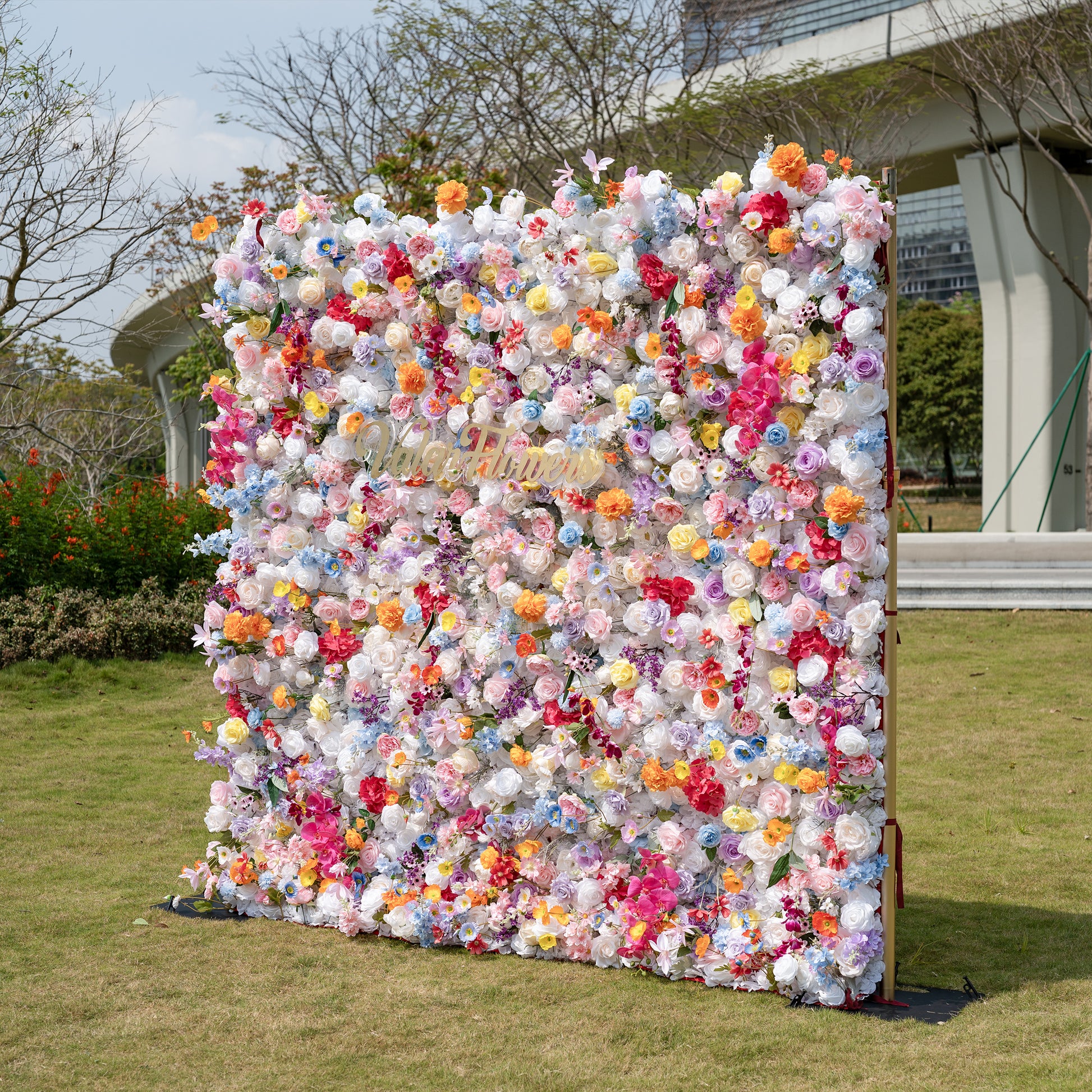 This is a showstopping, dense artificial flower wall backdrop from Valar Flowers, bursting with a vibrant, rainbow-hued mix of lifelike blooms. It features creamy white roses, soft pink peonies, sunny yellow poppies, vivid orange carnations, sky-blue delphiniums, deep magenta orchids, and delicate filler flowers, creating a lush, romantic focal point perfect for outdoor weddings, proposals, engagement parties, photo booths, and luxury event decor.