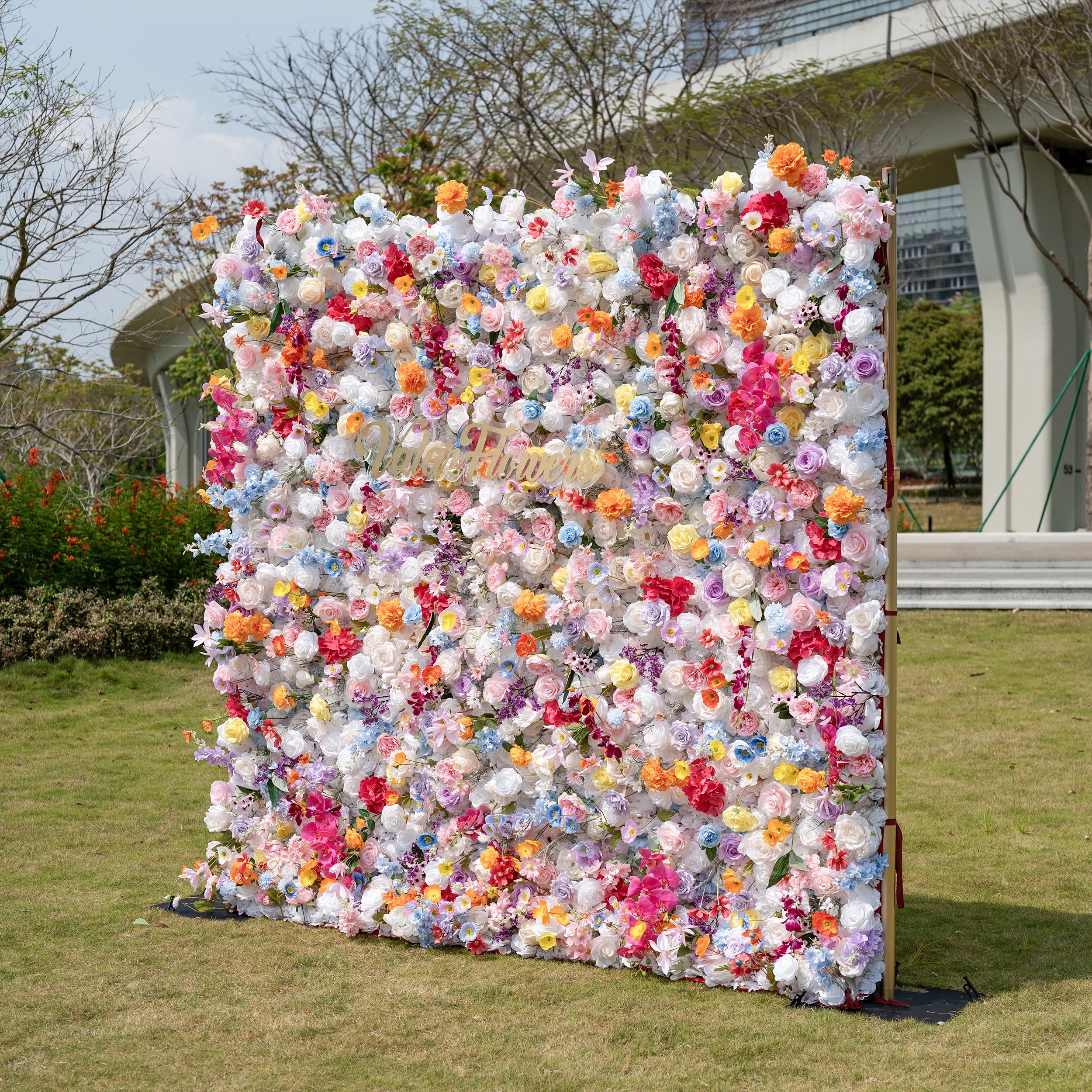 This is a showstopping, dense artificial flower wall backdrop from Valar Flowers, bursting with a vibrant, rainbow-hued mix of lifelike blooms. It features creamy white roses, soft pink peonies, sunny yellow poppies, vivid orange carnations, sky-blue delphiniums, deep magenta orchids, and delicate filler flowers, creating a lush, romantic focal point perfect for outdoor weddings, proposals, engagement parties, photo booths, and luxury event decor.